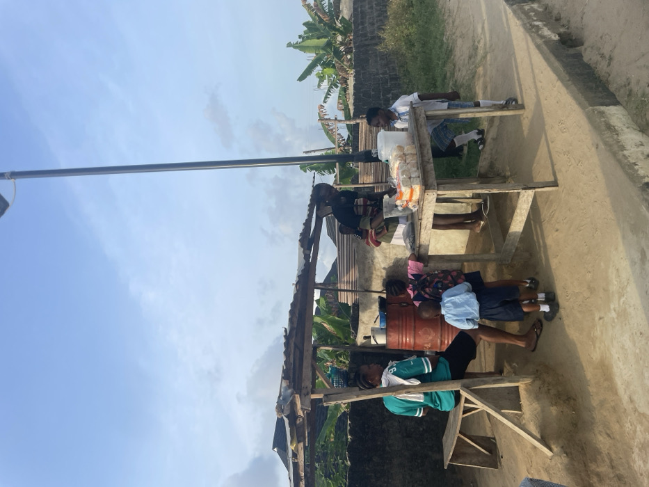 An outdoor market stall is depicted on a dirt path in Mosafejaw, Nigeria, under a light blue sky with scattered clouds. The stall consists of a rough wooden frame supporting a corrugated metal sheet roof. On the left, a woman in a teal shirt with a white cloth draped over her shoulder sits on a wooden bench. Adjacent to her is a red metal barrel topped with a cooking pot. A second woman, identifiable as a vendor, stands behind a wooden table displaying packaged bread, a clear plastic container, and a dark, unidentified pile. An infant is secured to her back. Three additional children are present: a boy in a light blue school uniform and dark shorts stands near the table, facing it; a girl in a pink outfit with a backpack stands slightly behind him; and a second boy in a white school shirt and plaid shorts stands to the right of the table, holding a white plastic pitcher. A tall, dark grey solar-powered streetlight pole is centrally located behind the stall. The background features a low concrete wall, a corrugated metal fence, and dense green foliage, including banana trees.