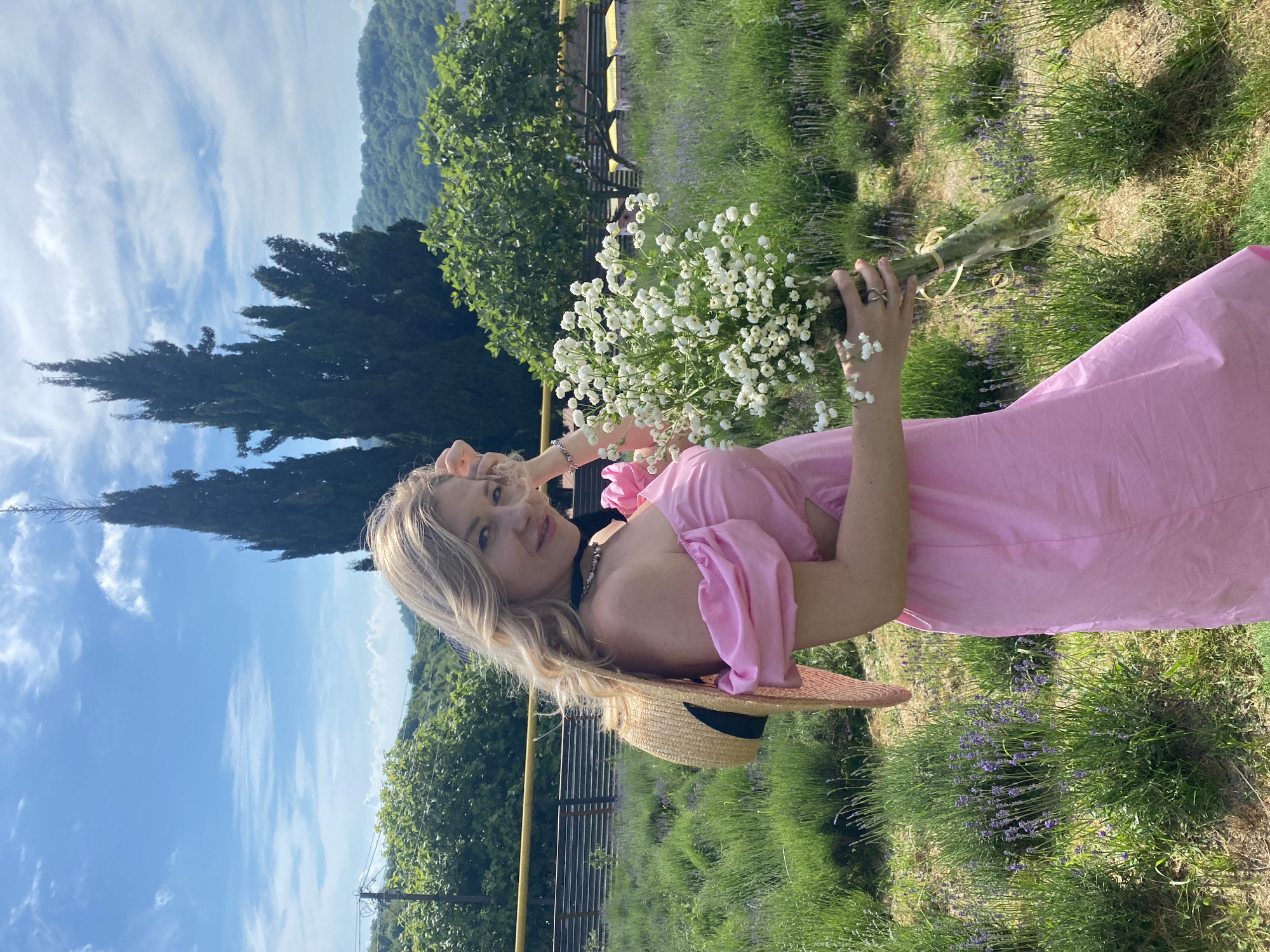 An adult Caucasian woman with blonde, wavy hair is standing in a lavender field in Verkhneye Uchdere, Russia. She is wearing an off-the-shoulder pink dress and a straw hat with a black ribbon, which is tilted against her back. The woman is holding a bouquet of small white flowers. In the background, there are tall green trees and a cloudy blue sky. The lavender field stretches out behind her, with rows of purple flowers and green foliage. The scene appears to be set on a sunny day.