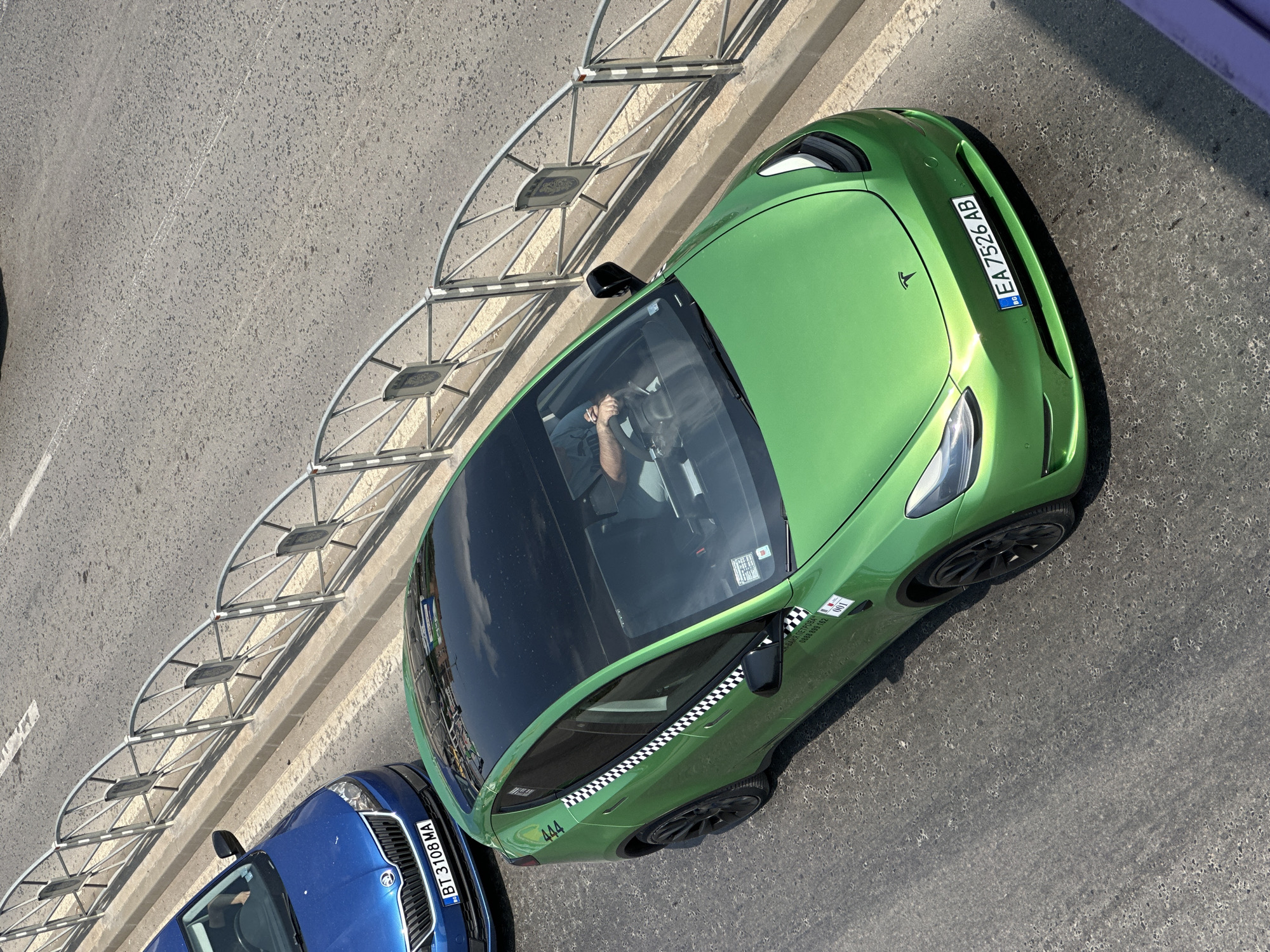 This is an aerial, eye-level shot of two cars on an asphalt road in Veliko Tarnovo, Bulgaria. The car in the foreground is a bright green Tesla Model Y with a black roof and black rims. It has a license plate that reads "EA 7526 AB" and the Tesla logo on the hood. The car has checkerboard detailing along the doors and a "444" decal on the rear side panel. There is a person driving the Tesla, with their hands on the steering wheel. The car behind the Tesla is a blue Skoda with a license plate reading "BT 3108 MA". Both cars are facing away from the camera, moving forward on the road. A metal railing with a decorative design separates the road from a concrete curb and sidewalk. The sun is casting shadows on the road, indicating it is daytime.