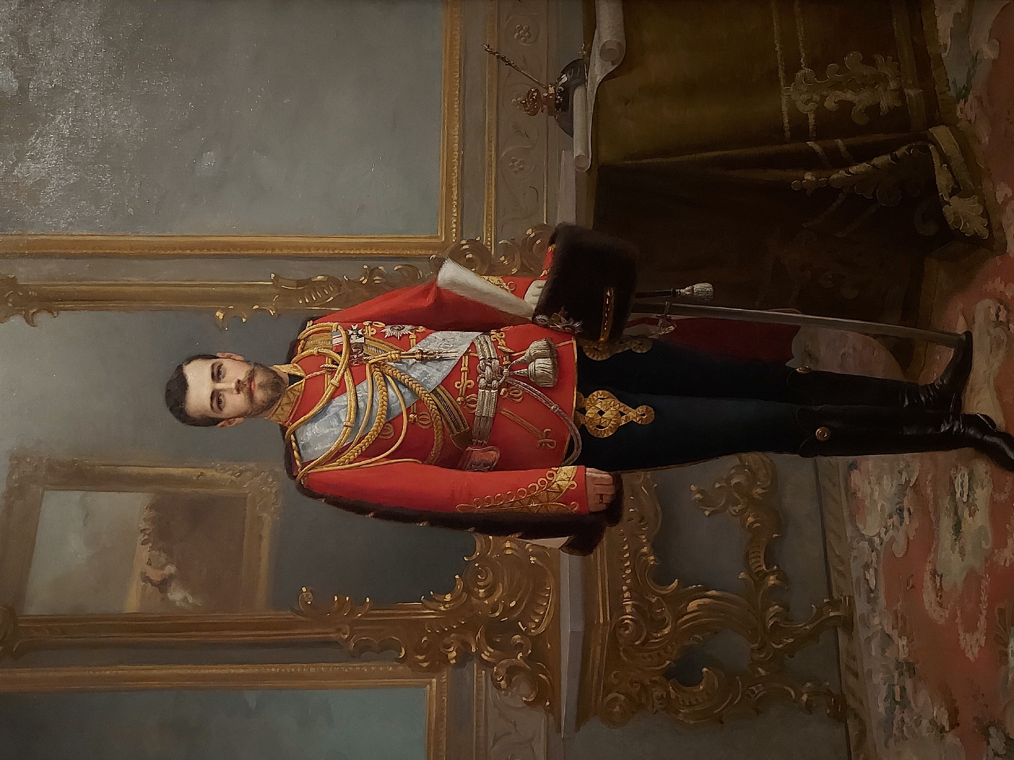 The image is a portrait painting of a man in a red military uniform with gold embellishments, standing indoors. He wears black boots and a dark fur hat is placed on a red cloth next to him. A sword lies next to the hat. He has dark hair and a trimmed beard. The background features ornate gold trim, a framed painting, a draped curtain, and a patterned rug on the floor. The portrait is likely located in Saint Petersburg, Russia, given the style and the subject's military attire which is characteristic of Russian Imperial portraits.