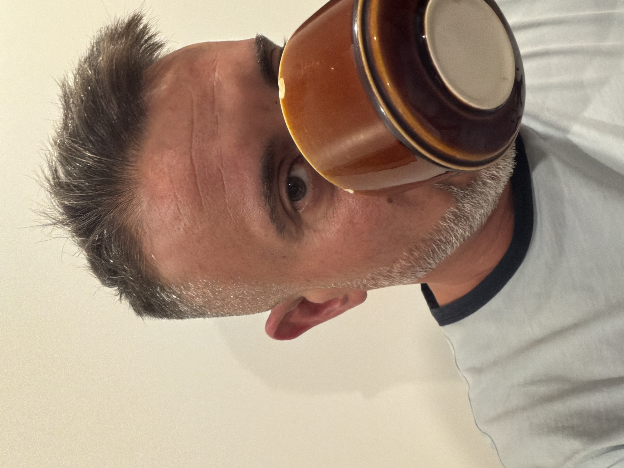 The image shows a close-up, waist-up view of a man drinking from a brown mug. The man has short, graying hair and a salt-and-pepper beard. He is wearing a light blue t-shirt with a dark blue collar. The background is a plain, light-colored wall. The man's left eye is visible, and he is holding the mug to his lips. The image does not provide any information about the location in Esch-sur-Alzette, Luxembourg.