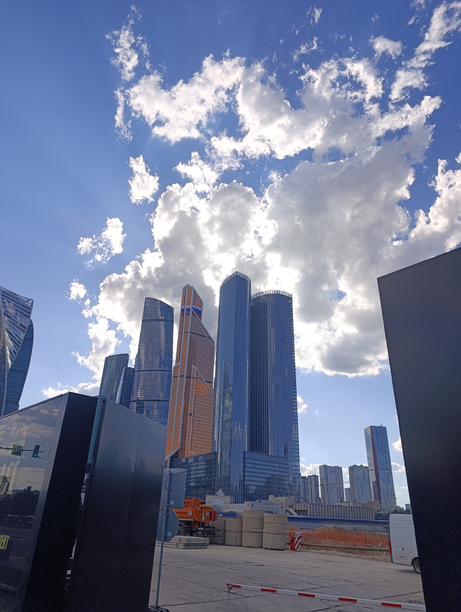 The image presents a modern urban landscape in Moscow, Russia, dominated by towering skyscrapers under a dynamic sky. The vibrant blue sky is filled with large, fluffy white cumulus clouds, with the sun partially obscured behind them, casting bright rays and illuminating the scene.
Numerous high-rise buildings form the city's skyline. On the left, a distinctive skyscraper with a twisted or undulating glass facade is visible. Toward the center, several sleek, dark blue/grey glass skyscrapers rise, reflecting the sky. One prominent building features sections with warm orange or bronze cladding, and near its peak, a pattern resembling the Russian flag (red, white, and blue stripes) is discernible. Further right, two very tall, cylindrical, blue glass towers stand close together, with other, slightly smaller buildings in the distance.
In the foreground and midground, the scene suggests an ongoing construction or development area. Two large, dark, rectangular structures, possibly art installations or temporary barriers, frame the view, with the left one reflecting the surrounding buildings and sky. Between these structures, a ground-level area features stacks of large concrete blocks, some red and white striped safety barriers, and an orange construction vehicle that appears to be a pump or mixer truck. A white utility van is partially visible on the right.
Visible text includes the partially obscured word "DENT" at the bottom of the left dark structure. On the orange construction vehicle, the word "МОБИЛЬНО" (MOBI'LNO, meaning "mobile" in Russian) is faintly visible.
The bright lighting indicates it is daytime, likely midday or early afternoon, with clear but cloudy weather. The modern architecture strongly identifies the location as the Moscow International Business Center, often referred to as Moscow City.