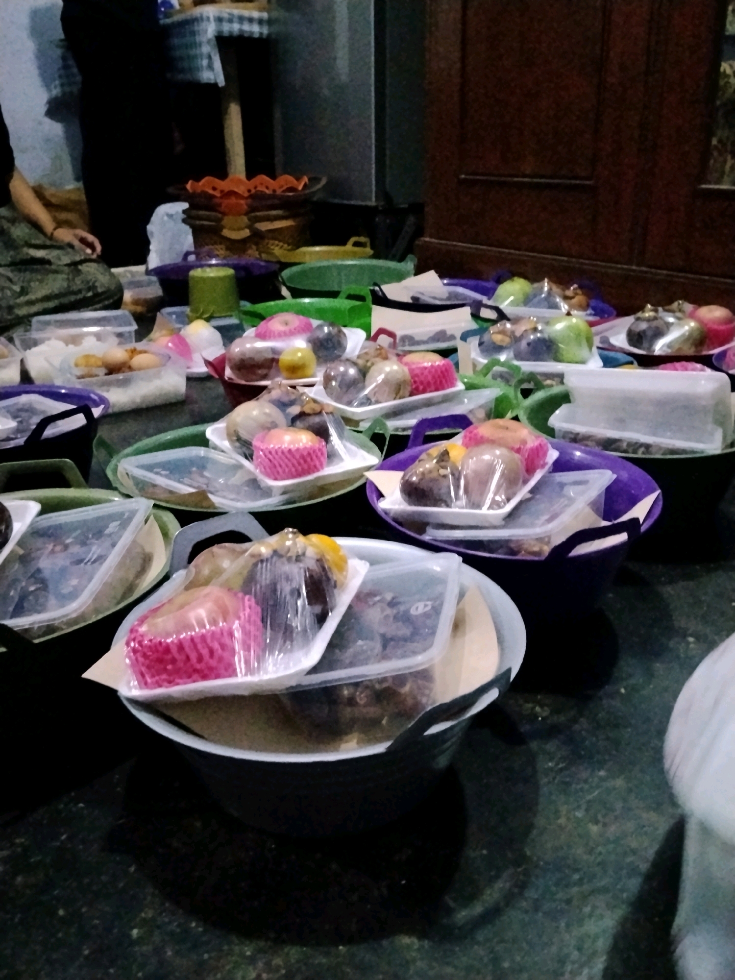 A dimly lit, indoor scene in what appears to be a home in Koncer Kidul, Indonesia, shows an abundance of pre-packaged food items, likely for an event. Several large, dark-colored plastic basins are arranged on a dark floor, each holding multiple white styrofoam trays filled with fruit. The fruit, including mangosteens, apples, and possibly other varieties, are individually wrapped in clear plastic. Some trays also contain pink paper cupcake liners, suggesting a mix of fruit and sweets. In the background, a person in dark clothing is partially visible, along with some baskets and wooden furniture. The overall impression is one of preparation and distribution of food.