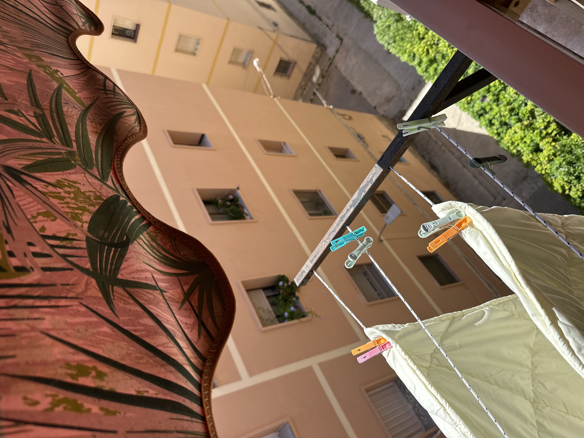 The image captures a low-angle view from a balcony, looking upwards and outwards. In the foreground, a section of a patterned awning with tropical leaf motifs is visible, draped and undulating. Below the awning, a clothesline is strung with what appears to be a light-colored blanket or duvet, secured by several colorful clothespins: pink, orange, and light green. The clothespins are gripping the fabric to the white rope.
Beyond the clothesline, the scene opens to reveal the facades of two residential buildings. The buildings are painted in a warm, peachy-orange hue, with lighter cream-colored horizontal banding. Multiple windows are visible on the building directly in view, some with small window boxes containing green plants. Further in the background, another building of similar color and style is partially visible, suggesting a dense urban environment. The ground level between the buildings shows a paved walkway and lush green foliage. The sky is bright, indicating a sunny day. The overall impression is one of domestic activity taking place in a Mediterranean or similarly warm climate. There are no people or animals visible in the frame.