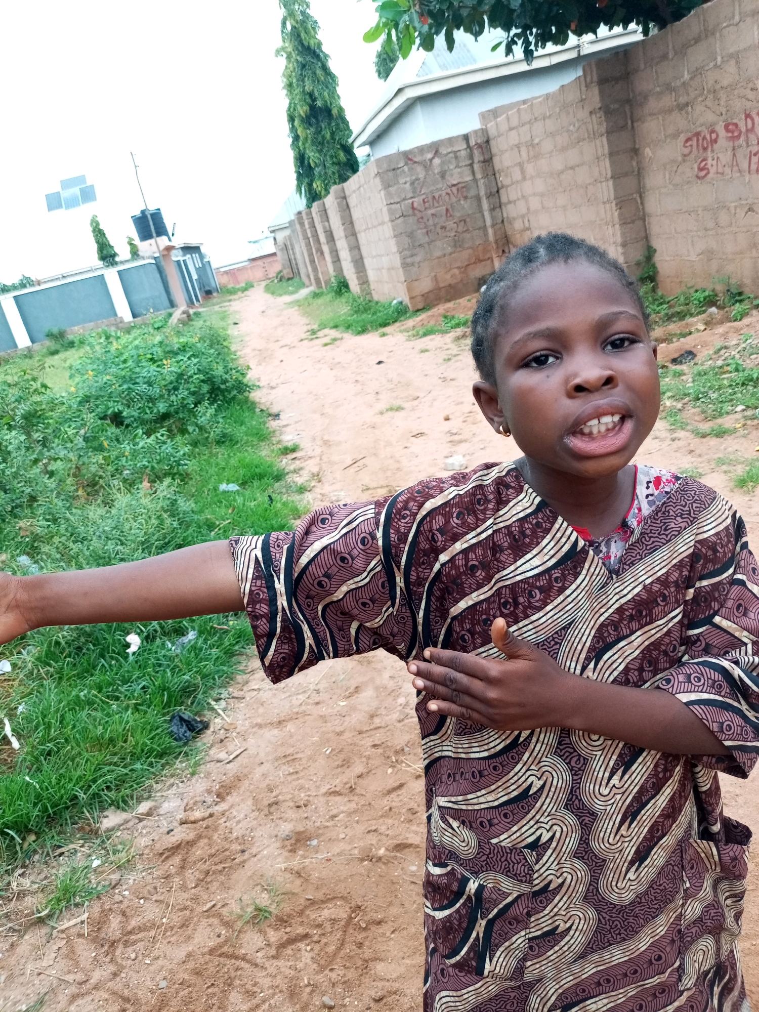 A young girl stands in an outdoor setting in Tempure, Nigeria. She is positioned in the foreground, looking towards the viewer with her mouth open and teeth visible. The girl wears a brown and cream patterned robe with pockets. Her left arm is extended outwards and slightly downwards, while her right arm is bent with her hand placed on her chest. Behind her, a dirt path runs diagonally across the frame, bordered by green grass and vegetation on the left. In the background, a brick wall with graffiti is visible, and further behind that, a building and trees are present. The overall lighting suggests an overcast day.