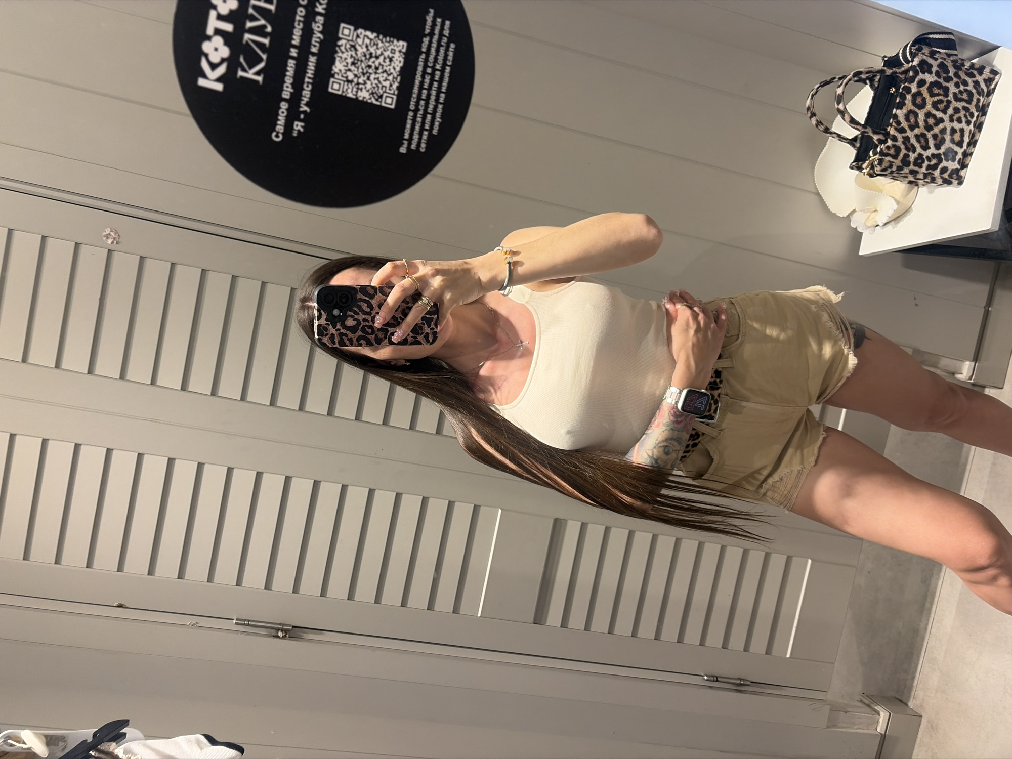 A young woman is standing in a fitting room, taking a selfie in a mirror. She has long, dark brown hair and is wearing a white tank top and light beige denim shorts. On her left wrist, she has a silver smartwatch with a leopard print band. She is also wearing several rings on her fingers and a silver necklace with a cross pendant.
The fitting room has light gray, slatted doors and walls. To the right of the woman, a circular black sticker with white text and a QR code is affixed to the wall. The text reads "КОТ klub" and "Самое время и место для...". Below the QR code, there is Russian text that translates to "You can scan the code to subscribe to us on social networks or go to Koton.ru for purchases on our website."
On a white shelf to the right, there is a small leopard print handbag and a white hat with a ribbon. The floor is tiled with light gray tiles. The overall lighting is bright and even, typical of a retail fitting room. The location is a Koton clothing store fitting room in Saint Petersburg, Russia, as indicated by the store's branding on the sticker.