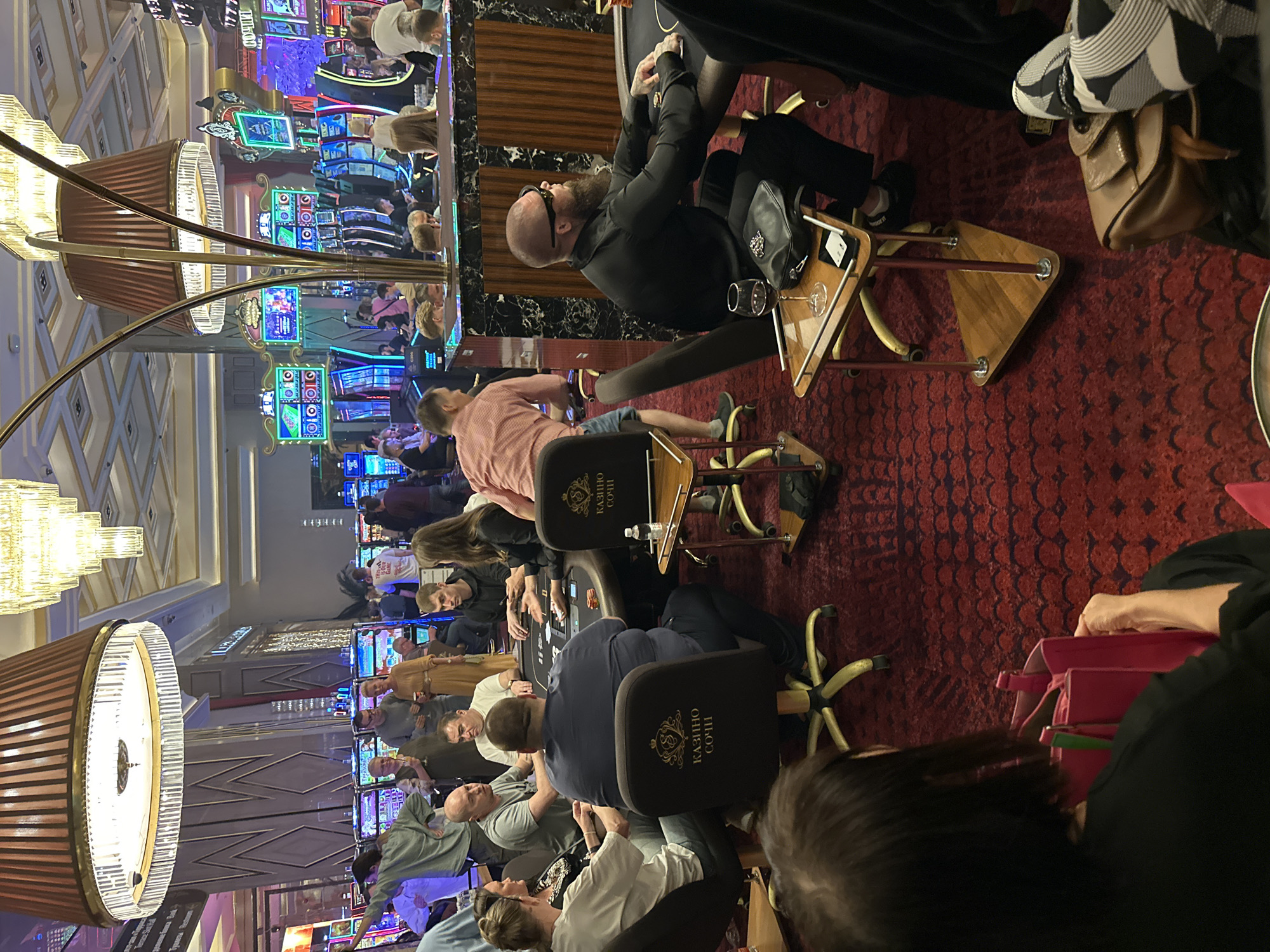 The image captures a scene inside a casino in Estosadok, Russia, likely part of the Olympic Park, a location known for its entertainment venues. The environment is dimly lit, with the primary illumination coming from overhead chandeliers and the vibrant, colorful lights of numerous slot machines that dominate the background. The flooring is a deep red carpet with a subtle pattern.
In the foreground, several individuals are seated around a card table, engaged in what appears to be a card game, possibly poker, indicated by the presence of chips and cards. One person, a heavy-set man in a dark blue shirt, is facing away from the camera, deeply engrossed in the game. Another man, wearing a black shirt and sunglasses with a prominent beard, is also seated at the table, looking to his right. A woman in a black jacket is to his right, her hands on the table, suggesting she is also participating. Other players are visible, their faces partially obscured or turned away.
To the left of the card table, a person in a white shirt is seated, their attention directed towards the game. Nearby, a person in a dark shirt is also present. A side table next to the man in the black shirt holds a glass of red wine, a black purse, and a mobile phone.
In the mid-ground and background, more people are milling about and playing at slot machines. The slot machines are brightly lit with various graphics and displays. People are seen standing and observing the games, and some are interacting with the machines. The overall atmosphere suggests a lively and potentially high-stakes gambling environment. The presence of multiple chandeliers, ornate lamps, and polished surfaces contributes to a sense of luxury and entertainment.