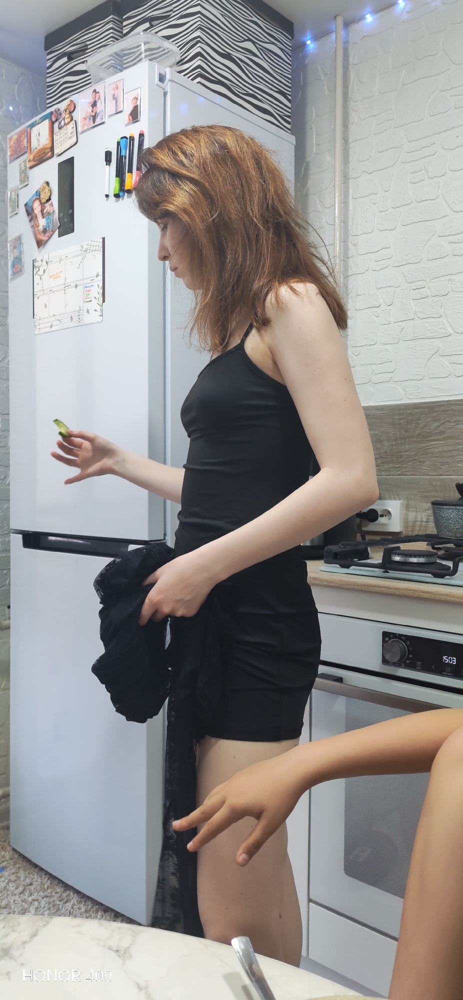A woman with reddish-brown hair stands in a kitchen in Noviy, Russia. She wears a black tank top dress and holds a piece of green produce in her left hand and a black lace garment in her right. A white refrigerator stands to her left, decorated with magnets, photographs, and a calendar. To her right is a white oven with a black stovetop. A child's arm and leg are visible in the foreground. The kitchen features a white brick-patterned wall and a ceiling with zebra-striped storage boxes. The image includes the "HONOR 400" watermark in the lower left corner.