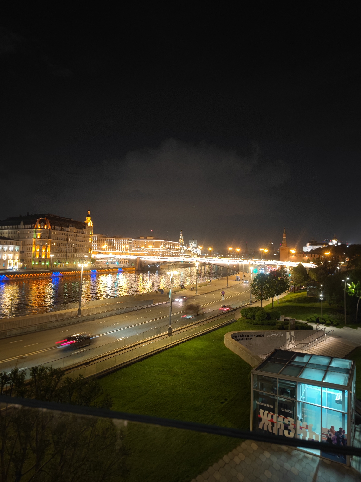The image depicts a nighttime view of a cityscape, likely a major city in Eastern Europe, given the architecture and language on display.
In the foreground, there's an enclosed structure with glass walls, potentially an entrance to an underground area or exhibition. The words "ЖИЗНЬ" (likely meaning "life" in Russian) are prominently displayed on the structure. Adjacent to this structure is a grassy area with a pathway leading down to the entrance, possibly for an underground exhibition area.
Beyond the immediate foreground, there's a road with cars moving, evident from the light streaks. Further out, a river reflects the lights from the buildings on the opposite bank. Buildings are illuminated, some with ornate architecture and towers. A bridge spans the river, heavily illuminated with lights that reflect in the water.
In the distance, the Kremlin and surrounding structures are visible, further indicating that this scene is likely in Moscow, Russia.
The sky is dark, with some clouds obscuring the view. The overall scene is well-lit, creating a contrast between the dark sky and the illuminated cityscape.