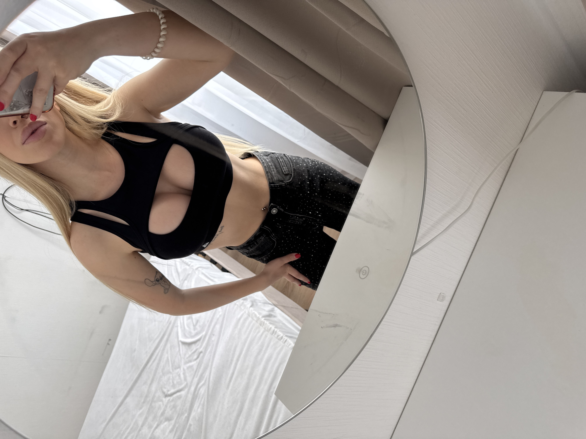 A woman is taking a mirror selfie in what appears to be a bedroom. She is wearing a black crop top with a cutout at the chest and dark, sparkling jeans. Her blonde hair is visible, and she is holding a phone in her right hand. The reflection in the mirror shows a white bed with white sheets in the background. To the right of the woman, there are beige curtains. The overall setting is a domestic environment. Petro-Slavyanka is a village in Russia, not a city.