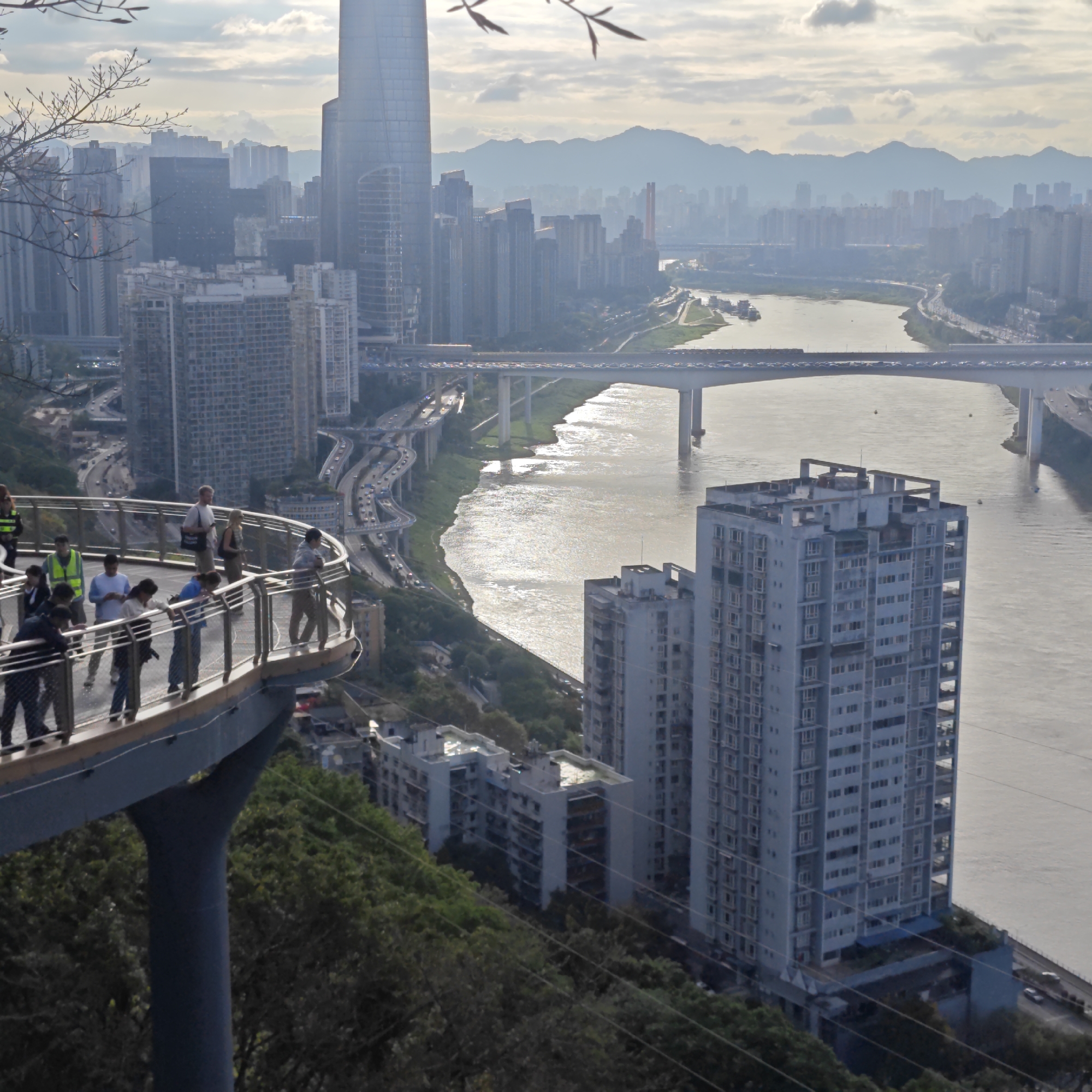 This expansive image presents a bustling cityscape in Yuzhong District, China, viewed from a high vantage point, likely an observation deck. In the foreground, a curved, elevated viewing platform hosts several people. Dressed casually, they are seen leaning on the railings, observing the panoramic urban sprawl and the river below. Their activity suggests they are tourists or sightseers taking in the scenery.
The scene is dominated by a dense array of modern architecture, featuring numerous high-rise residential buildings and towering skyscrapers. A wide, light-colored river flows through the city, its surface shimmering with reflections under a soft, possibly late afternoon or early morning light. Spanning the river is a substantial, multi-lane bridge, heavily trafficked with vehicles. Below and alongside this main bridge, a complex system of elevated highways and roads winds its way through the mountainous and hilly terrain, also carrying moving traffic.
Distant mountains, partially obscured by a light atmospheric haze, form the backdrop to the cityscape, blending into a partly cloudy sky. Along the riverbanks and on the hillsides, patches of green trees and foliage are visible, interspersed with urban development. A few boats or barges can be spotted on the river. Bare tree branches in the upper left corner subtly frame the dynamic urban vista. The overall impression is of a thriving, modern metropolis seamlessly integrated into its natural, rugged landscape. No discernible text is visible within the image.