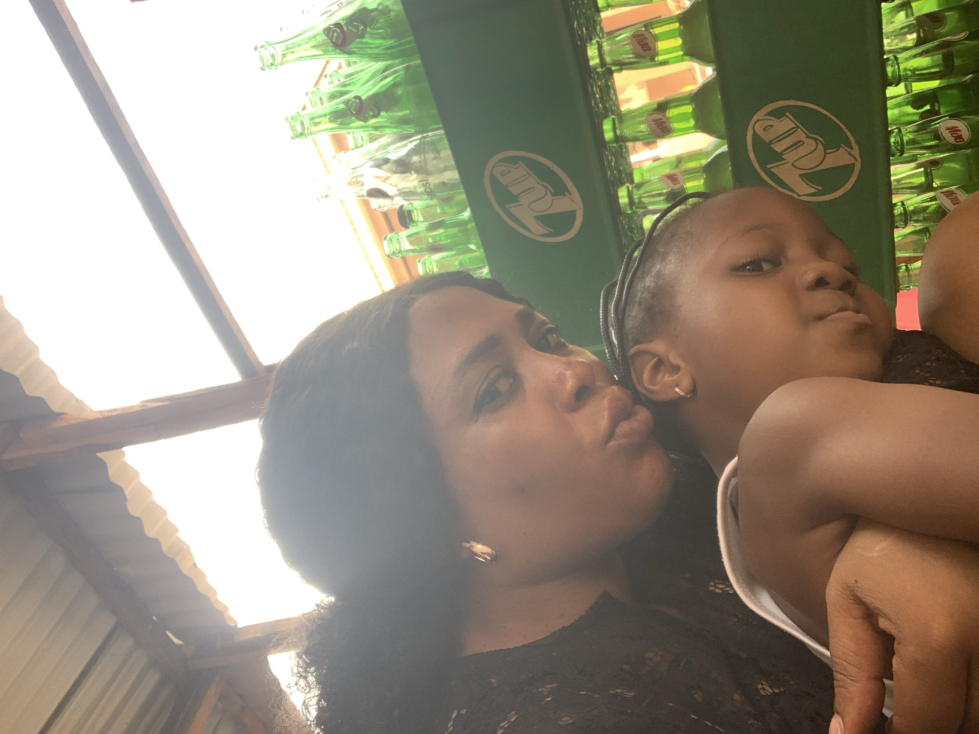 The image shows a woman and a child in what appears to be a semi-outdoor structure in Sabon Tasha, Nigeria. The woman has dark curly hair, is wearing a black lacy top and earrings, and is making a kissing face. The child, with braided hair, is wearing a white tank top. Above them is a display of green glass bottles labeled "7up". The roof above is made of corrugated metal sheets.