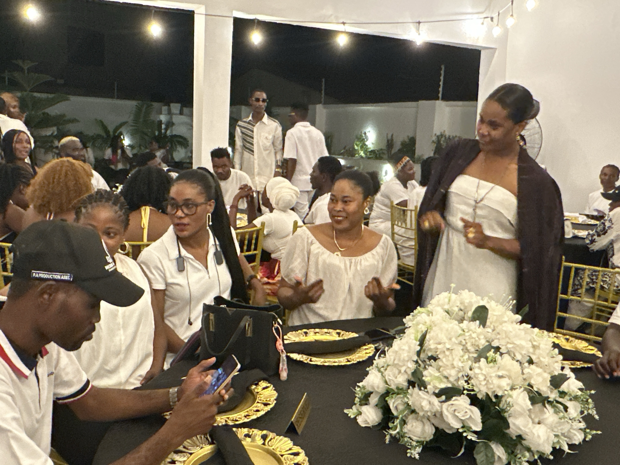 This media file depicts an outdoor evening event in Barden Yana, Nigeria. A group of people are gathered around black tables adorned with gold-rimmed plates and white floral centerpieces. Many attendees are wearing white attire, suggesting a formal occasion or a dress code.
In the foreground, a person wearing a black cap and a white polo shirt with red and blue trim is looking at their phone. Behind them, a woman in a white off-the-shoulder top with a dark jacket draped over her shoulders is interacting with someone out of frame. Her expression suggests animation and engagement. Other individuals are visible in the mid-ground and background, seated at tables or standing, some conversing.
The scene is lit by string lights overhead, indicating it is nighttime. The background shows a building with white pillars and railings, and vegetation suggesting a garden or outdoor event space. The presence of multiple tables and a gathering of people indicates a social event, possibly a celebration, party, or reception. The overall atmosphere appears lively and social. The cap worn by the person in the foreground has text on it, reading "P.A.PRODUCTION ASST".