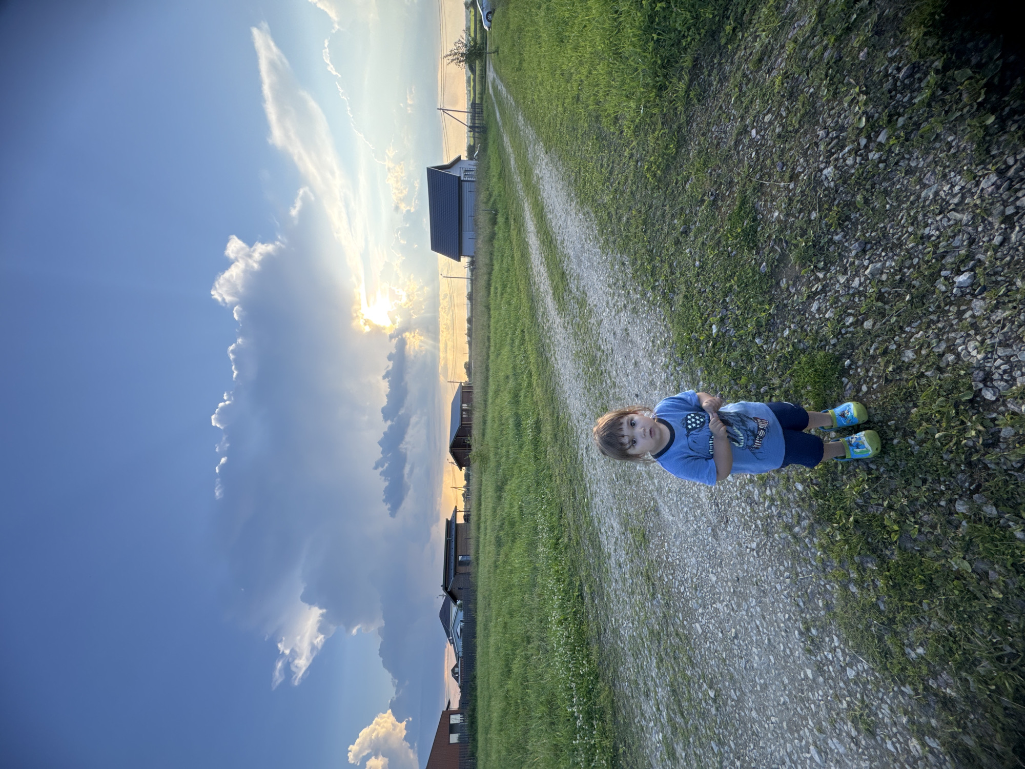 A young child stands on a gravel path in a grassy field, with several houses visible in the background under a dramatic sky filled with clouds and the setting sun. The child is wearing a blue t-shirt and dark shorts, and colorful shoes. The path leads away from the viewer into the distance. The scene appears to be a rural area, consistent with the outskirts of Kempelevo, Russia, characterized by open fields and scattered residential buildings. The time of day is likely late afternoon or early evening, indicated by the low angle of the sun and the quality of light.