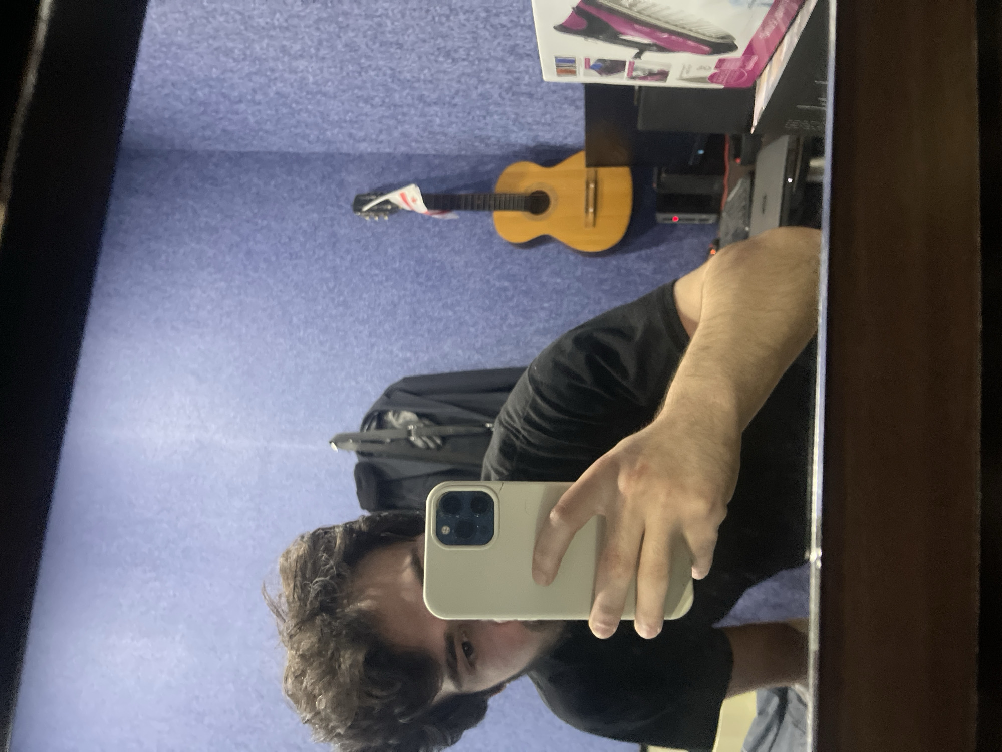 The image is a reflection in a mirror of a person holding a smartphone. The person is wearing a black t-shirt and has dark, curly hair. The background is a purple, textured wall. On the wall, a guitar is hanging, adorned with a white and red flag. To the right of the guitar, there is a black object, possibly a computer monitor or speaker. In the foreground, a shelf or desk is visible, with some electronic devices and a pink and white box. The mirror itself has a dark frame, and the reflection shows the lower part of the room, including what appears to be a bed with blue bedding on the left side of the image. The overall impression is of a personal space, possibly a bedroom or studio. There are no specific visual cues that definitively place this scene in Tbilisi, Georgia.