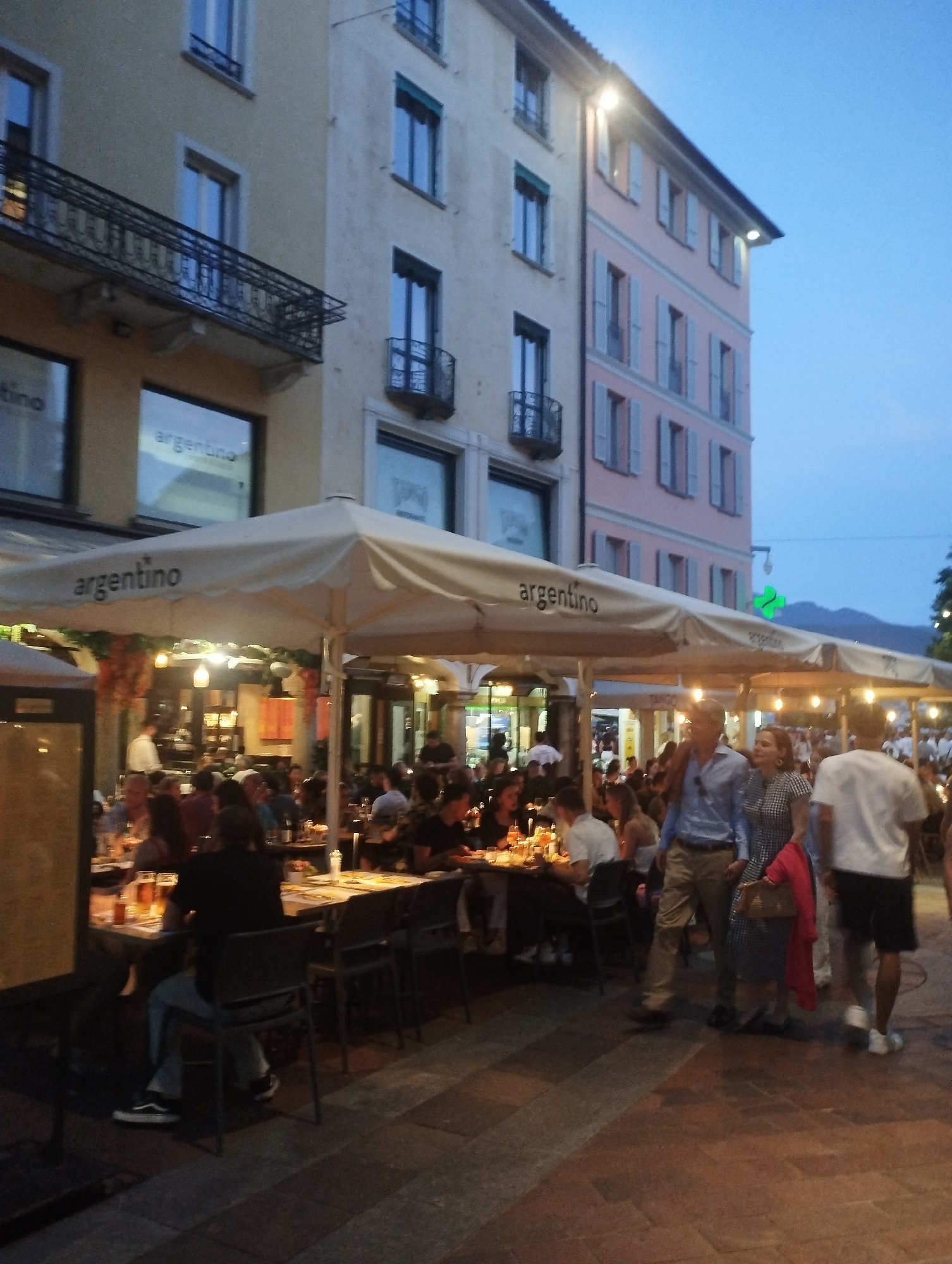 The image depicts an outdoor dining scene in Lugano, Switzerland. Several people are seated at tables under large white umbrellas, enjoying a meal. The umbrellas are branded with the word "argentino". In the background, there are buildings with windows and balconies. The sky is a dusky blue, suggesting it is either early evening or late afternoon. The lighting appears to be artificial, with string lights illuminating the dining area. A few individuals are walking through the scene, while others are seated, engaged in conversation or eating. The ground is paved with cobblestones.