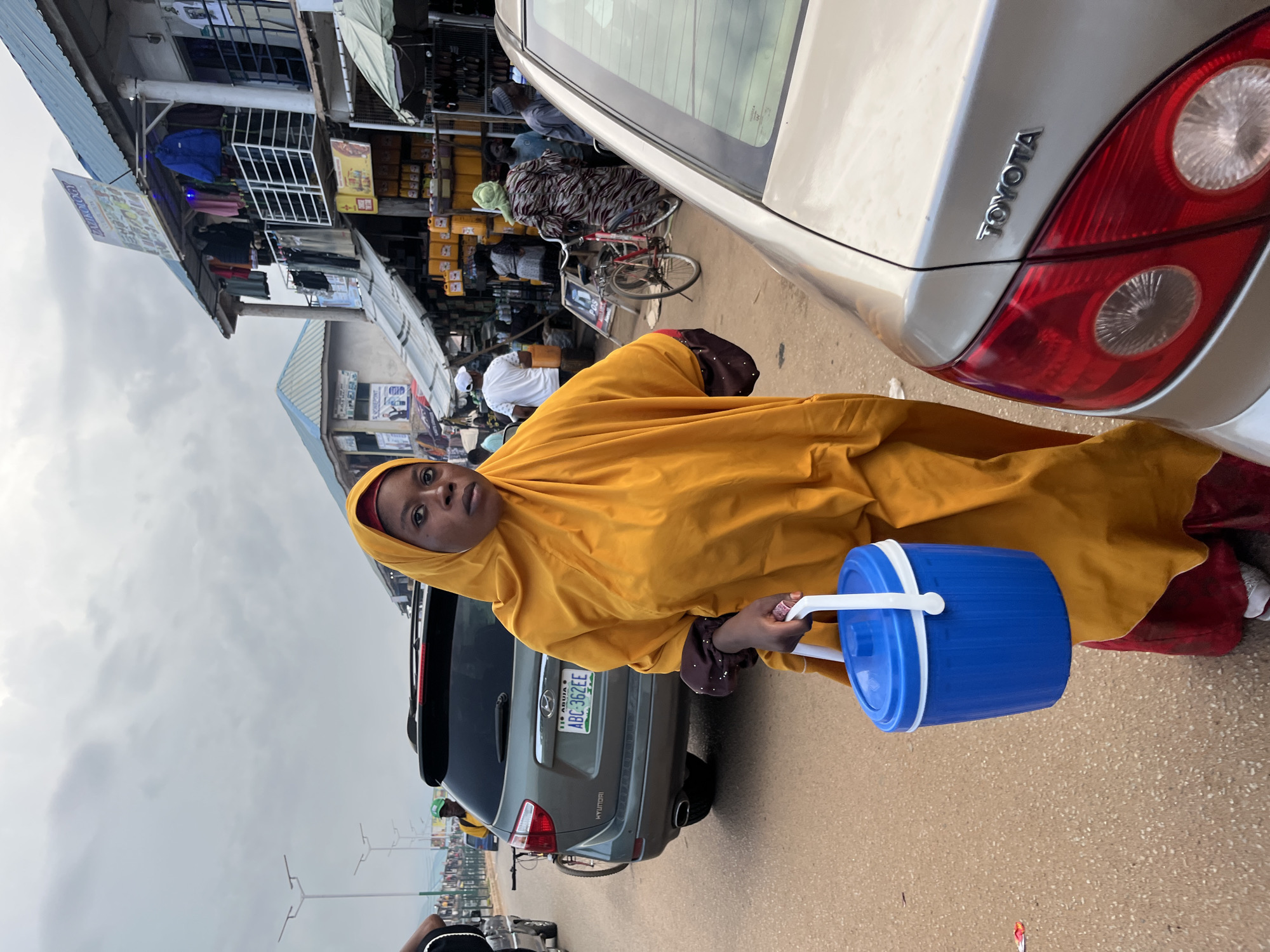 A young woman wearing a yellow hijab and traditional attire stands in a street market, holding a blue and white water cooler. She is positioned next to a silver Toyota sedan. In the background, stalls line the street, displaying clothing and various goods. Several people are visible, some browsing at the stalls and others walking through the market. A grey Hyundai SUV is parked behind the Toyota. The scene suggests a busy day in a Nigerian market, likely in the morning or afternoon given the lighting. The sky is overcast, indicating cloudy weather. The license plate on the Hyundai reads "ABC 362EE" and "FCT ABUJA". The woman's expression is neutral, and she appears to be looking towards the camera. The overall atmosphere is one of everyday commerce in a bustling urban environment.