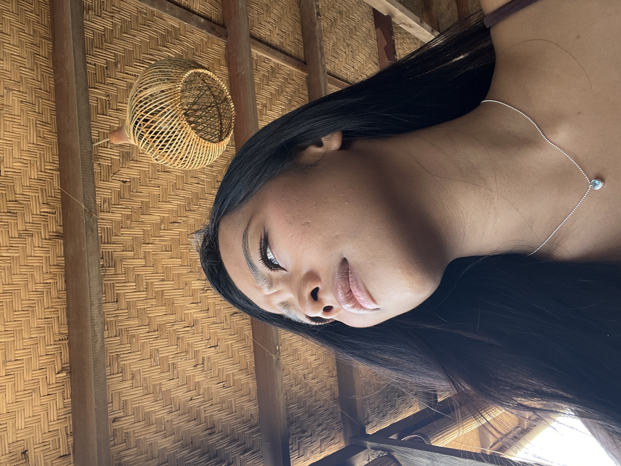 A low-angle, medium shot shows a young woman with dark hair and a silver necklace with a blue heart pendant looking up and to her left. She is wearing a dark colored top. Her face is partially obscured by her hair. The background is a thatched roof with wooden beams and a woven light fixture hanging from the ceiling. The lighting suggests it is daytime. The image appears to have been taken indoors, possibly in a resort or restaurant in North Kuta, Indonesia, due to the rustic architectural style.