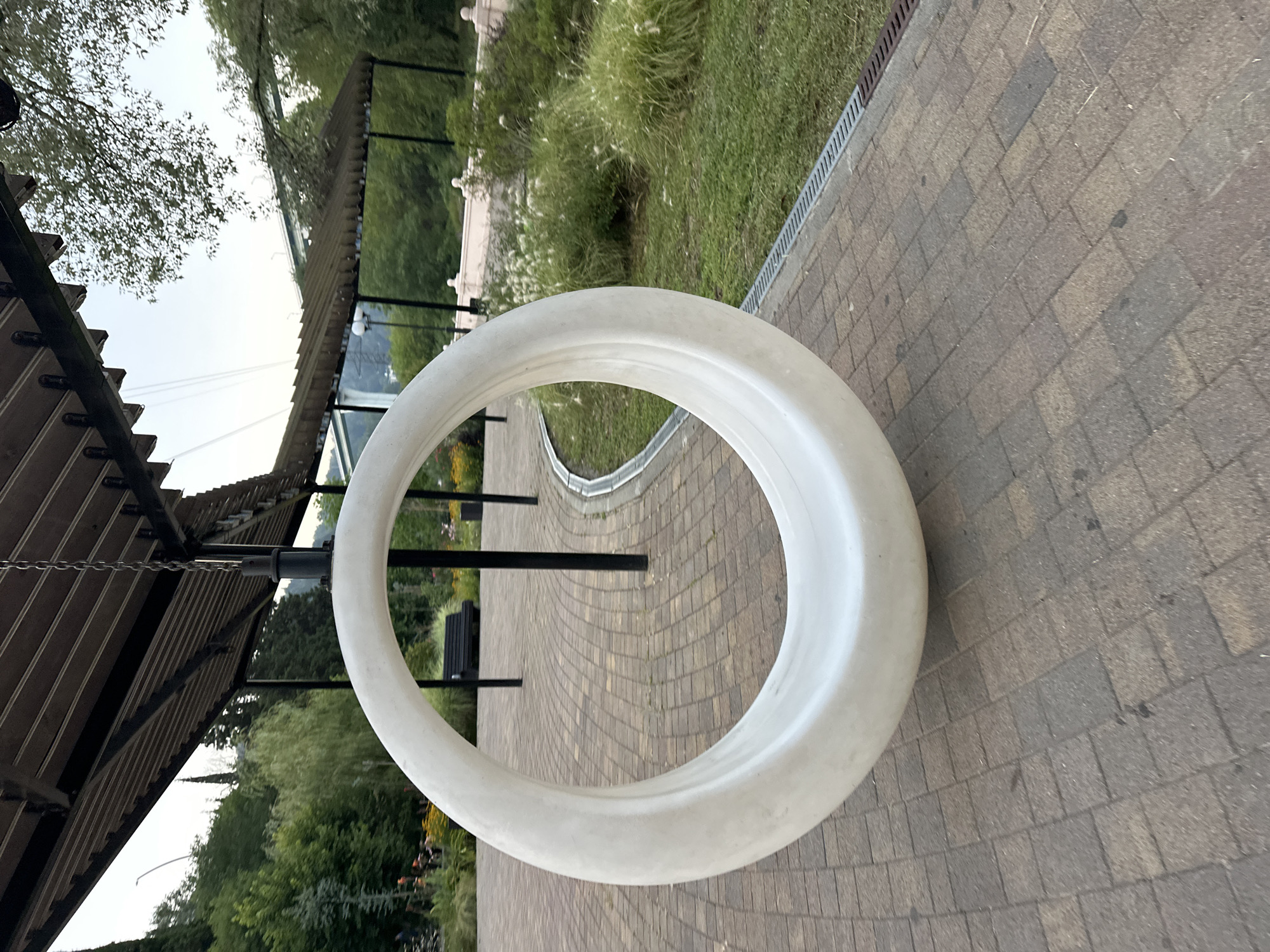 An off-center, low-angle shot captures a large, white, circular swing hanging from a dark, slatted roof structure. The swing is the primary subject and is positioned in the foreground, appearing smooth and solid. The ground beneath is paved with interlocking tan and gray bricks arranged in a herringbone pattern. To the right of the swing, a patch of green grass is visible, bordered by a narrow metal drain grate. In the background, lush green trees and some flowering plants line the area. A dark bench is visible in the mid-ground, set against a backdrop of trees and foliage. Beyond that, a large bridge with multiple spans can be seen in the distance, with a hazy, overcast sky above. The overall scene suggests an outdoor public space, possibly a park or promenade, in Sochi, Russia, given the architectural style of the roof and the presence of lush vegetation common in the region.