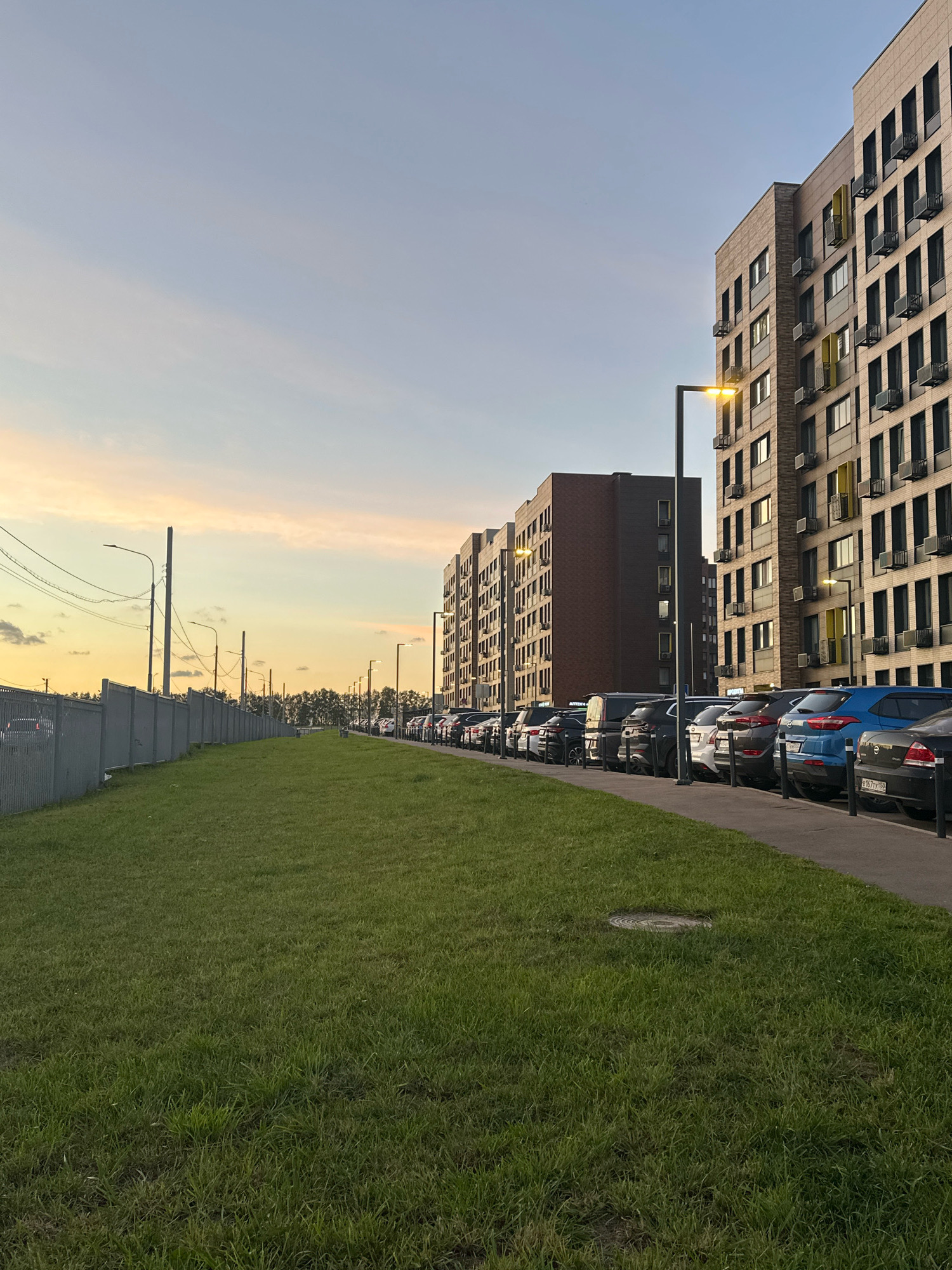This is a landscape photograph taken at dusk in the city of Vidnoye, Russia. In the foreground, a strip of green grass slopes gently upwards towards a paved area. A manhole cover is visible within the grass. To the right, a row of parked cars lines the street, with the rear ends of several vehicles facing the camera. The cars are of various colors, including black, blue, and silver. Beyond the cars, two modern, multi-story apartment buildings stand, their facades predominantly brick-toned with yellow accents around some windows and air conditioning units. A street lamp illuminates the scene. To the left of the grass, a grey metal fence runs along the edge of the property, with utility poles and wires visible behind it. The sky above is a soft gradient of pale blue and yellow, with wispy clouds. The overall impression is a quiet residential street scene at the end of the day.