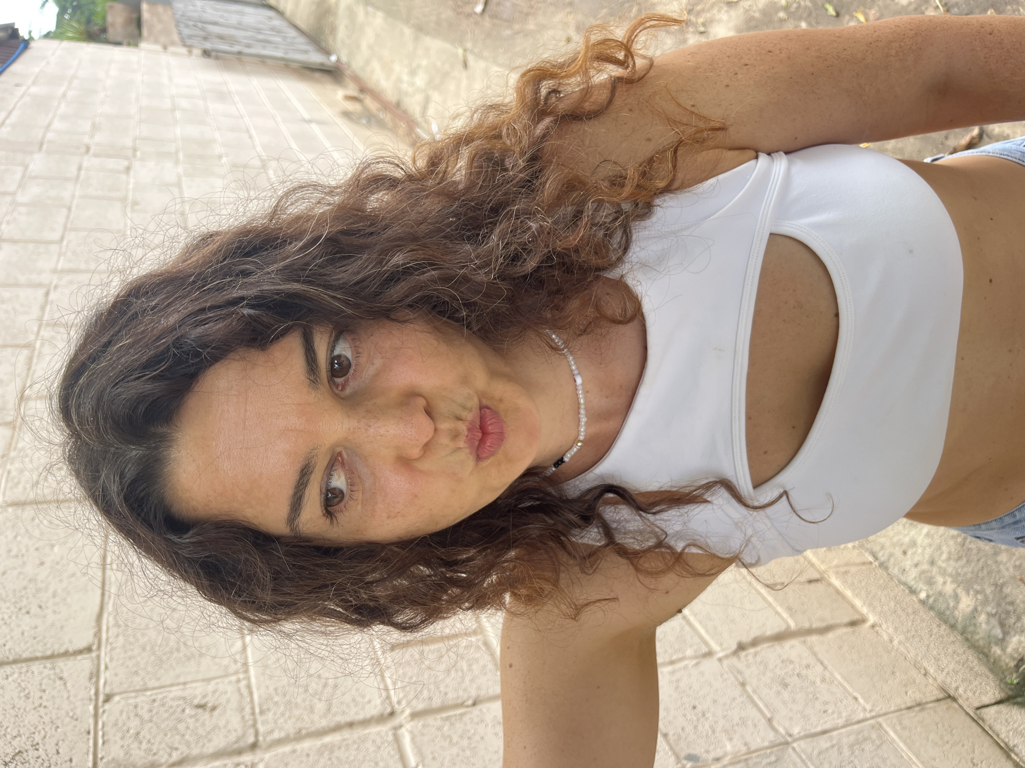 A woman with long, curly brown hair is standing outdoors, leaning against a light-colored brick wall. She is looking to her left and puckering her lips. She is wearing a white crop top with a cutout in the center and a beaded necklace. She also appears to be wearing denim shorts, though only a small portion of them is visible. The background consists of the brick wall on the left and a concrete walkway or street on the right, with a fence and some greenery visible in the distance. The lighting suggests it is daytime. The image is a medium shot, focusing on the woman's upper body and face.