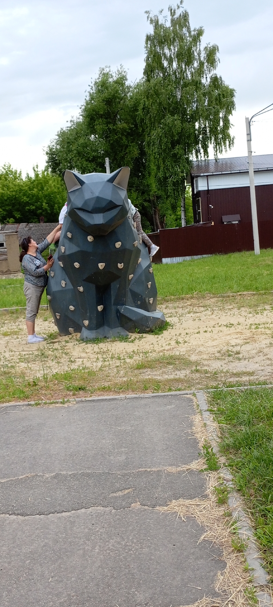 In Kirsanov, Russia, on a cloudy day, a dark grey, polygonal cat-shaped climbing structure sits on a sand-covered area. The structure has multiple rock-climbing holds on its surface. A woman in a patterned long-sleeved shirt and light-colored capri pants stands to the left of the cat structure, her hand reaching towards it. A child, partially visible, is climbing on the right side of the cat structure. In the background, trees with lush green foliage are present, along with a brown building and a white pole. A paved path with scattered dry grass runs along the bottom of the image.