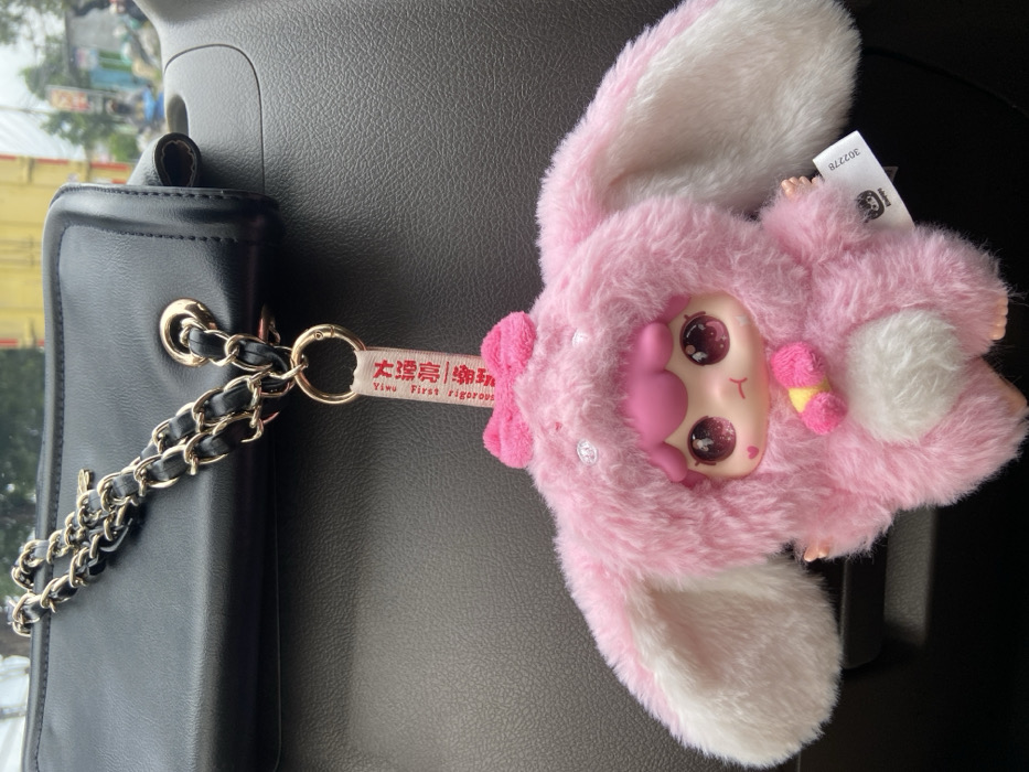 A black purse with a gold chain rests on a gray surface. Attached to the purse is a pink, fuzzy keychain featuring a character with large eyes and rabbit ears. The keychain has a tag with text in what appears to be Chinese and English. The plush keychain appears to have a fluffy tail and a small, pink and yellow accessory attached to it.