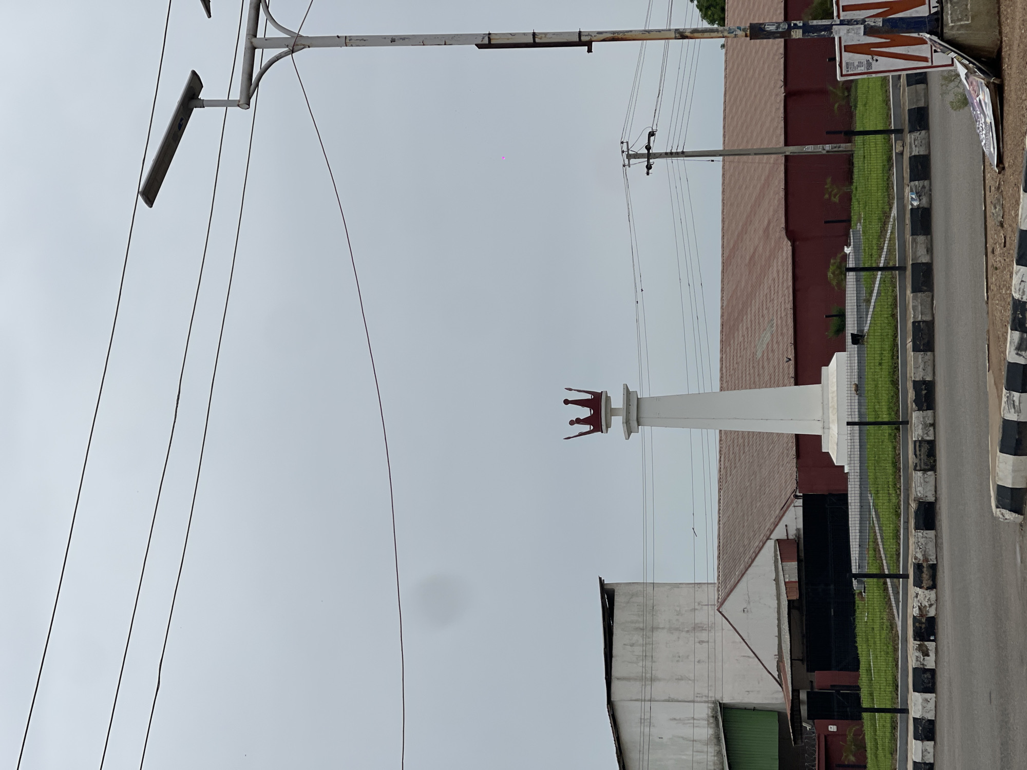 The image depicts a street scene in Jos, Nigeria. A white pillar with a red crown on top stands prominently in the center of the frame. The sky is overcast and gray. Overhead power lines crisscross the view. A light fixture is attached to a pole on the left side of the image. A building with a red roof and a black fence is visible to the right of the pillar. A road with black and white striped curbing is also present on the right. Another building, appearing aged with a white wall and green section, is on the left. A partially visible sign is present on the right.