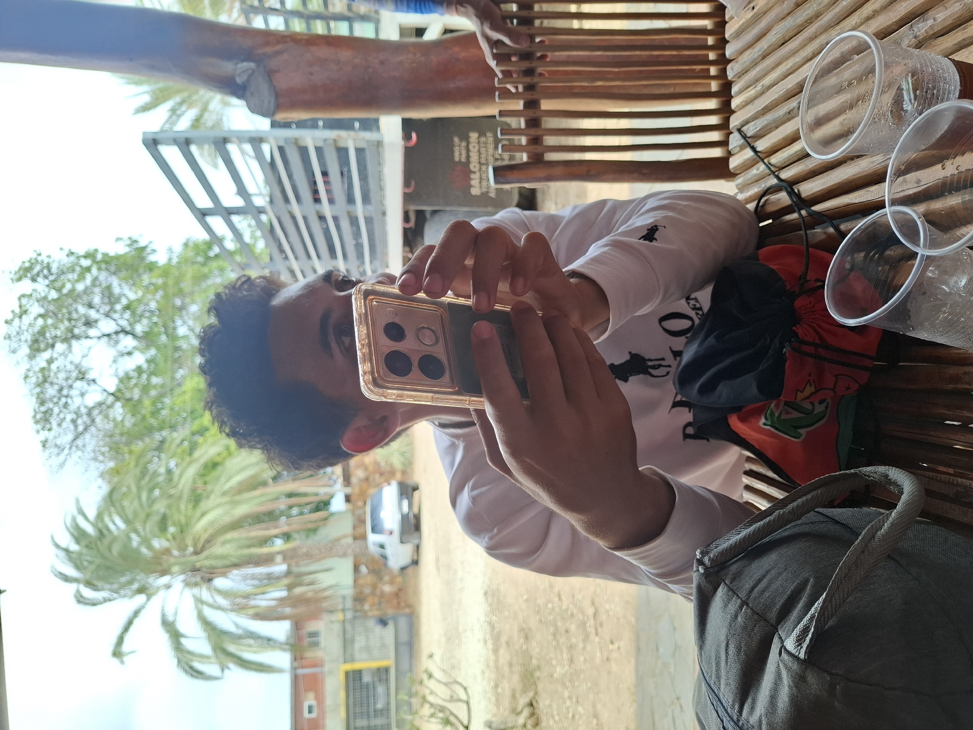 A man sits at a wooden slat table outdoors, holding a smartphone with a clear case in front of his face. His curly hair is dark. He is wearing a white, long-sleeved shirt. On the table are several clear plastic cups and a red and black drawstring bag. A gray backpack sits nearby. Beyond the table, a view includes palm trees, buildings, and a white vehicle. A sign for "SALOMON TRUCK PARTS" is visible. The setting suggests an urban or suburban environment with trees and buildings. The exact location in the city of Unknown, Unknown is unidentifiable.