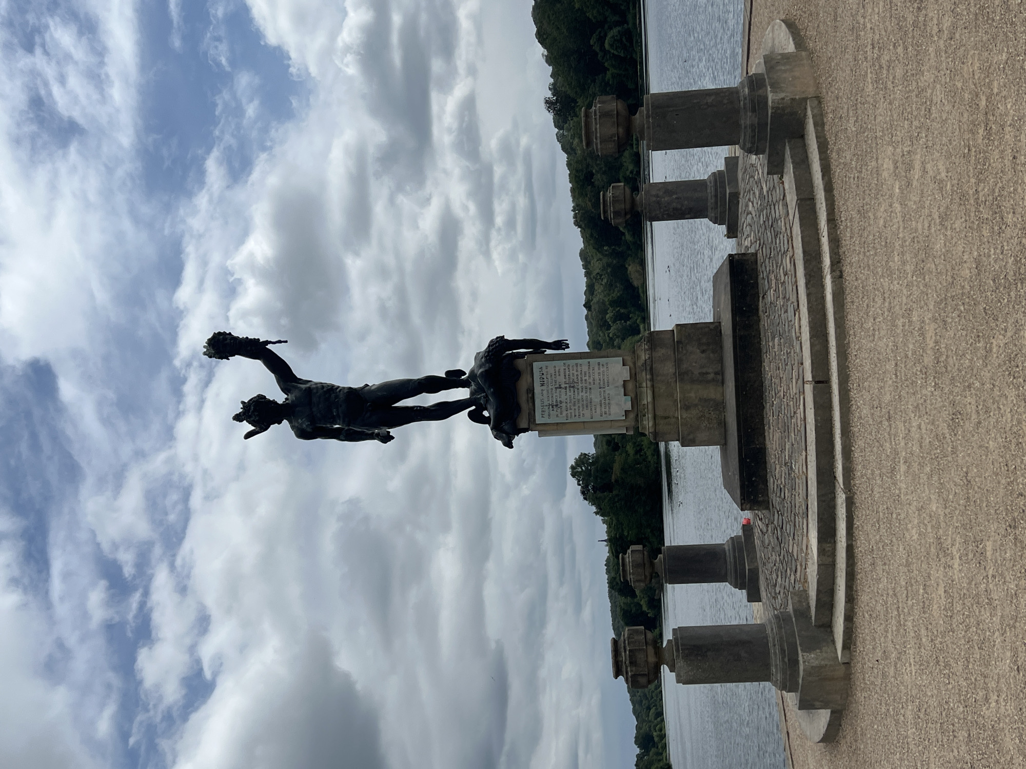 This is a vertical-format, outdoor, eye-level shot featuring a dark bronze statue of Perseus holding Medusa's head. The statue is positioned in front of a body of water and a line of trees. The sky above is filled with dramatic white and gray clouds. The statue stands on a pedestal which has a plaque with text. The plaque reads: "PERSEUS AND MEDUSA. ORIGINAL STATUE SCULPTED BY RIVINITO CELLINI IN FLENCE FOR THE GRAND DUKT OF TUSCANY MOL RIPUKA MARE GEDIE OF DUKE ONS THERLAND WITH THE FLEMISSION FRIEND THE THEN CLAND DUKE OF TUSCANY WAS ORIGINAL ALLY FRICCIO MERT ON THE ONSTRUCTION OF TRENTHAM MDCCCXL HERE RESTORED AND REPLACED HU BY KUZABETH CORINTESSO SUTHERLAND". The statue is part of a garden setting, with stone pillars and paving stones visible in the foreground. The overall impression is one of historical significance and grandeur, set against a somewhat overcast sky. The time of day appears to be daytime, and the weather is cloudy. The setting suggests a park or garden with a prominent water feature.