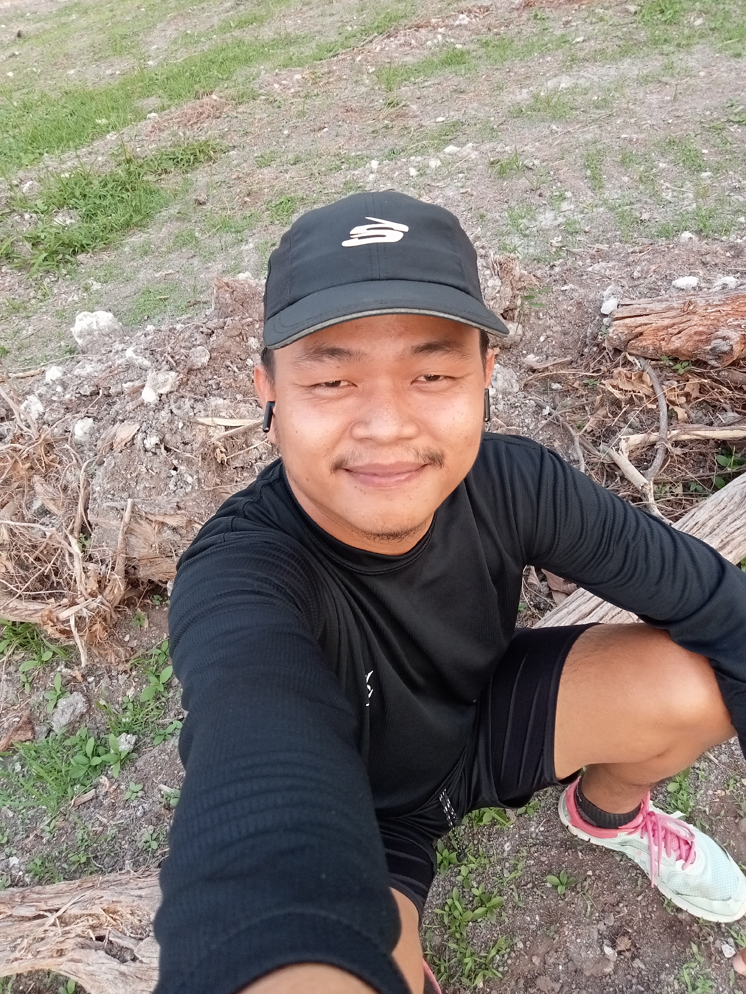 A man wearing a black baseball cap with a white logo, a black long-sleeved shirt, black shorts, and white and pink running shoes is sitting on the ground outdoors. He is wearing black earbuds. The ground is covered in dirt, rocks, and dry vegetation, with some green grass visible. There are also several pieces of fallen wood. The scene appears to be a rural or undeveloped area, potentially on the outskirts of Teluk Kenidai, Indonesia, given the arid terrain and sparse vegetation.
