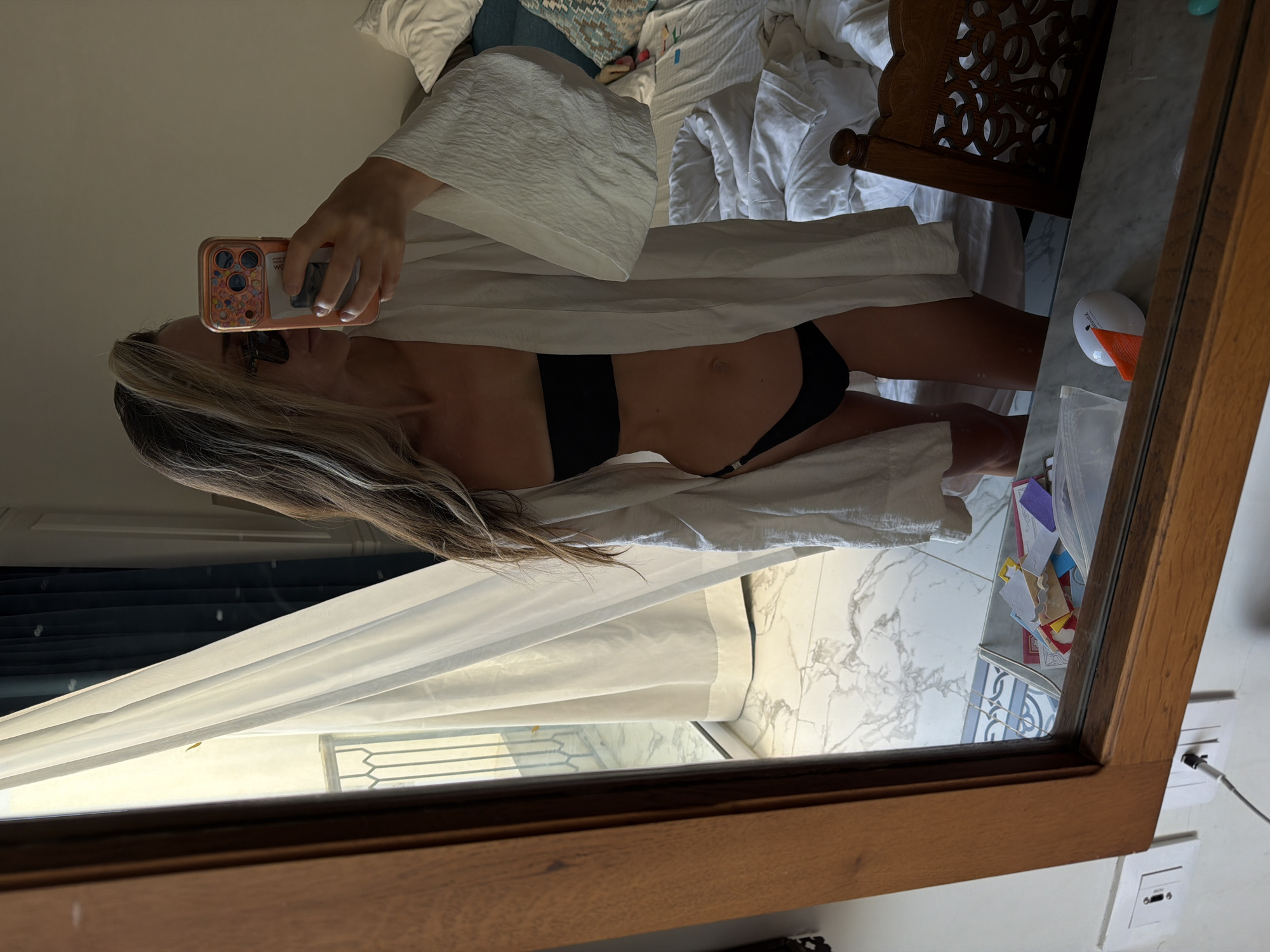 This mirror selfie captures a woman in a bright, indoor setting, likely a bedroom or hotel room in Sharm El Sheikh, Egypt. The image is oriented horizontally, showing the reflection of the woman standing upright.
The subject is a woman with long, blonde and brown hair, wearing a black bikini top and matching bottoms. She is partially covered by a white, textured, open robe or bathrobe. She holds a smartphone with a decorative case in her right hand, taking the mirror selfie. Sunglasses are visible on her face or forehead.
Behind the woman's reflection, a bed with white, rumpled bedding and pillows is visible, along with a carved wooden headboard. To her left, a sheer white curtain filters bright natural light coming from a window, suggesting it is daytime. A dark window blind and what appears to be a balcony railing are faintly visible beyond the curtain.
To the right in the reflection, on what seems to be a vanity or dresser, a small white object with an orange cap is visible, possibly a cosmetic device or diffuser. Next to it, a container holds various colorful paper cutouts or craft items. The floor visible in the reflection features a white and grey marble pattern. Below the wooden mirror frame on the wall, two white power outlets are present, one with a charging cable plugged in. The scene conveys a relaxed, casual atmosphere, typical of a vacation or personal space.