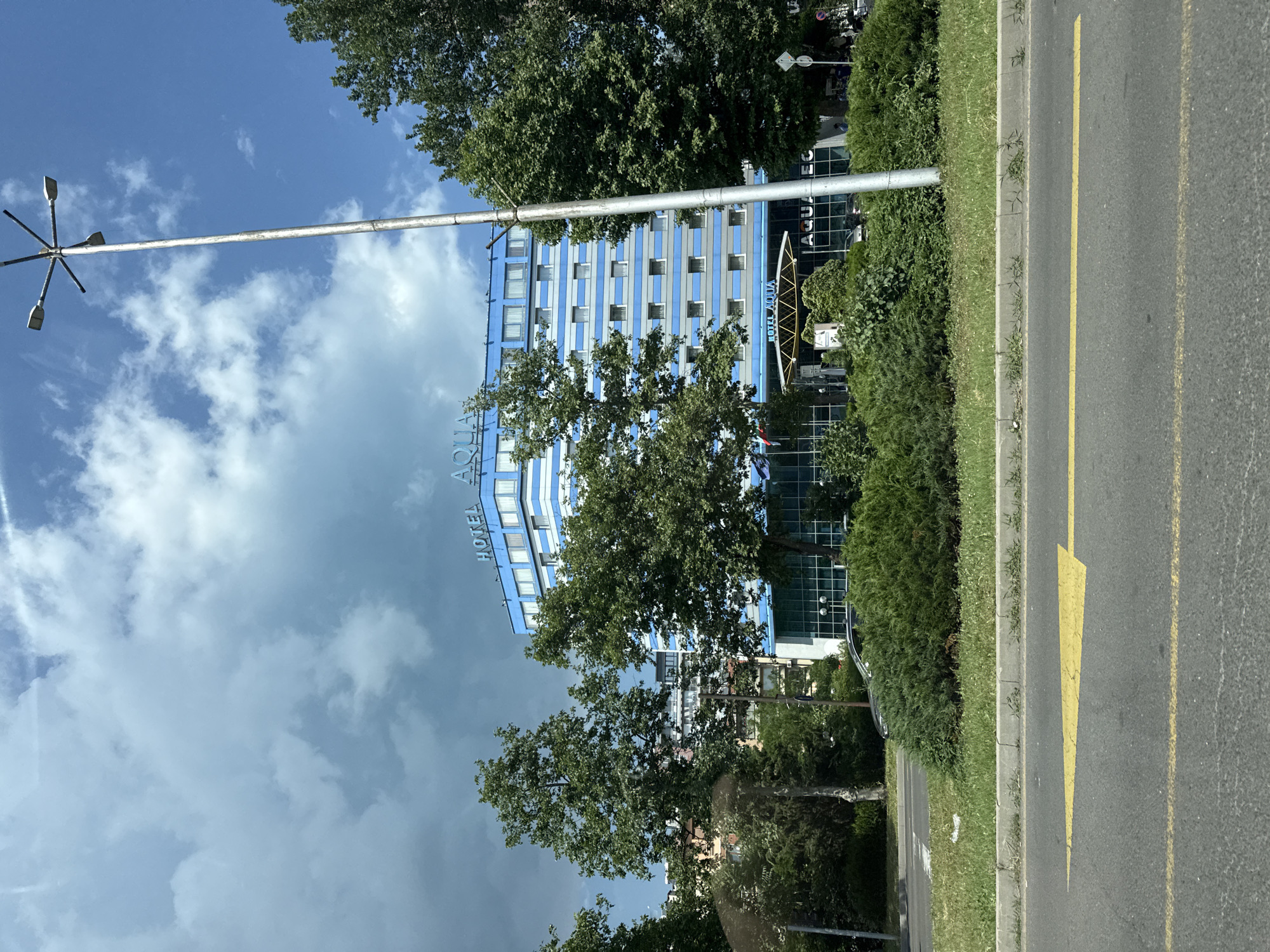 The image shows the Hotel Aqua in Burgas, Bulgaria. It is a multi-story blue and white striped building with "HOTEL AQUA" signage at the top. The building is surrounded by green trees and bushes. In the foreground, there is a road with yellow directional arrows painted on it and a concrete curb. A tall lamppost with multiple lights stands to the right of the hotel. The sky is partly cloudy with a mix of blue sky and dark clouds. The scene appears to be a daytime shot, possibly during a change in weather.