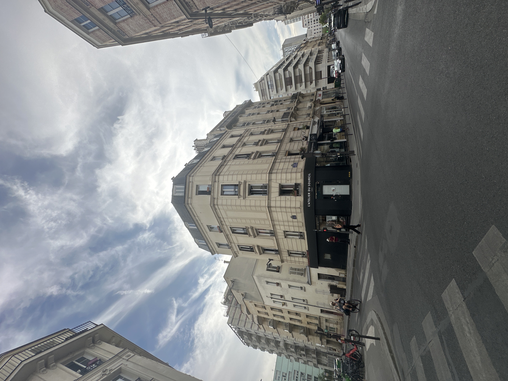 The image depicts a street corner in Paris, France, with several multi-story buildings lining the road. A prominent beige building with a dark storefront occupies the center of the frame. The storefront has signage that reads "L'ATELIER DU SOURCEIL PARIS". A pedestrian crosses the street at a marked crosswalk. Several bicycles are parked on the left side of the street, and two people are on a bicycle further down the sidewalk. Cars are driving on the street, and the sky above is filled with wispy clouds. The overall scene is an urban streetscape with a blend of architectural styles, indicative of Parisian city life.