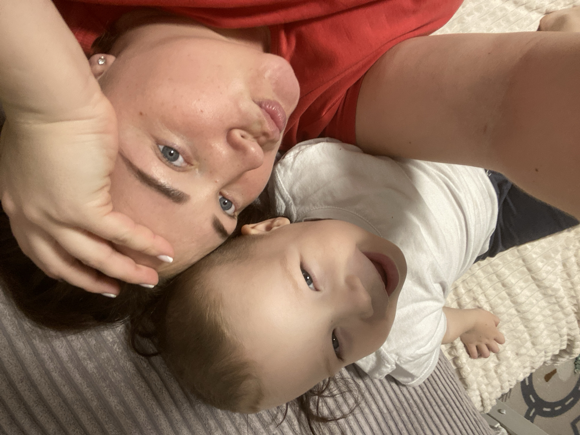 A selfie shows a woman and a baby lying on a bed. The woman has light skin, blue eyes, and dark eyebrows. She is wearing a red shirt. Her left hand is touching her forehead. The baby is wearing a white shirt. The bed is covered with a grey and beige blanket. The location is not discernible from the image and cannot be determined as Sochi, Russia.
