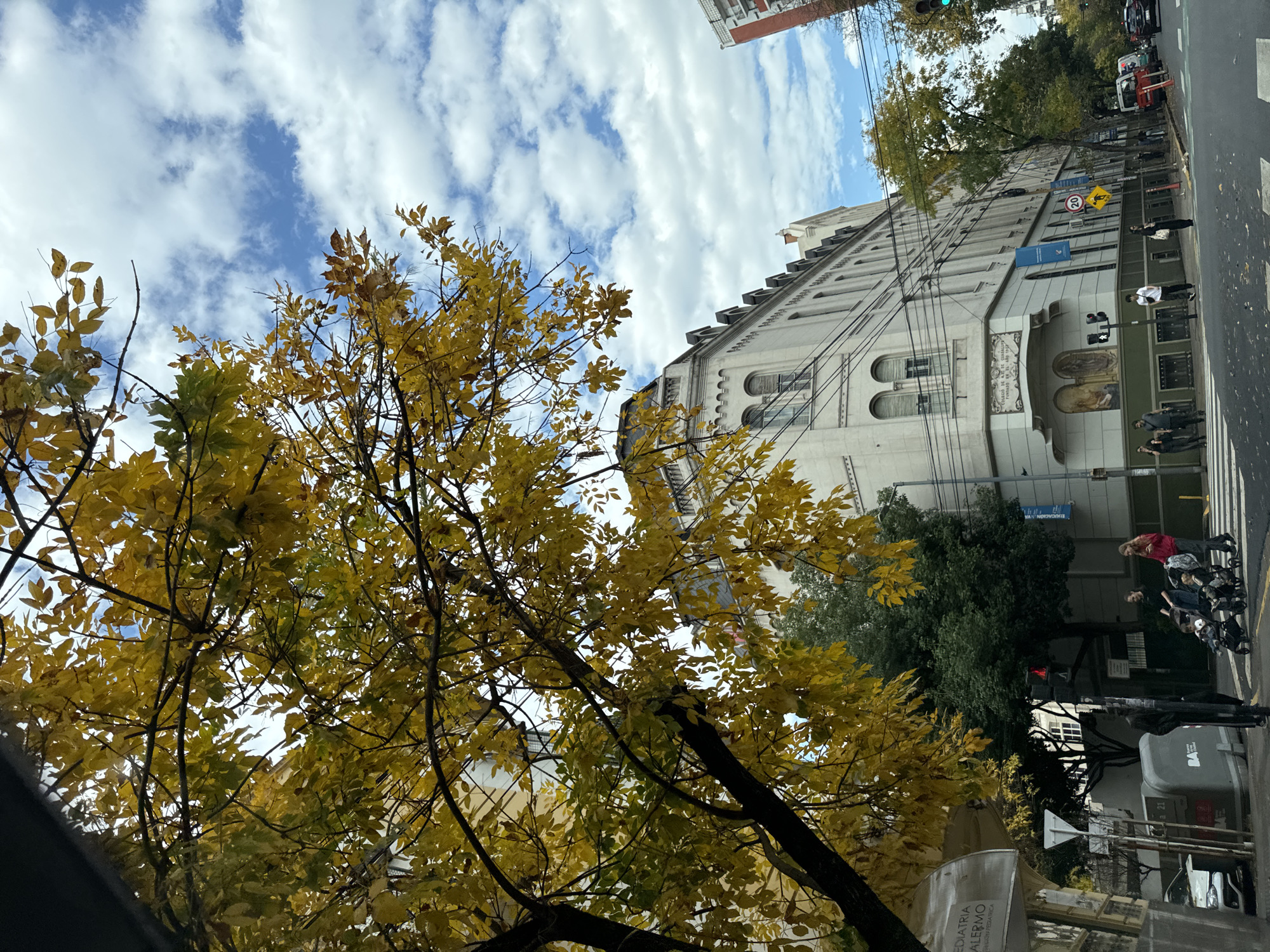 The image captures an autumn scene in Buenos Aires, Argentina. Dominating the foreground is a tree with yellow leaves, characteristic of the season. Behind the tree, a light-colored building with a gabled roof and arched windows stands prominently. Its facade features architectural details and possibly murals above the entrance.
The setting appears to be a city street. Pedestrians are visible on the sidewalk in front of the building, along with parked cars and a traffic light. A "20" speed limit sign is visible, indicating a slow-speed zone. The sky is filled with puffy white clouds against a backdrop of blue.