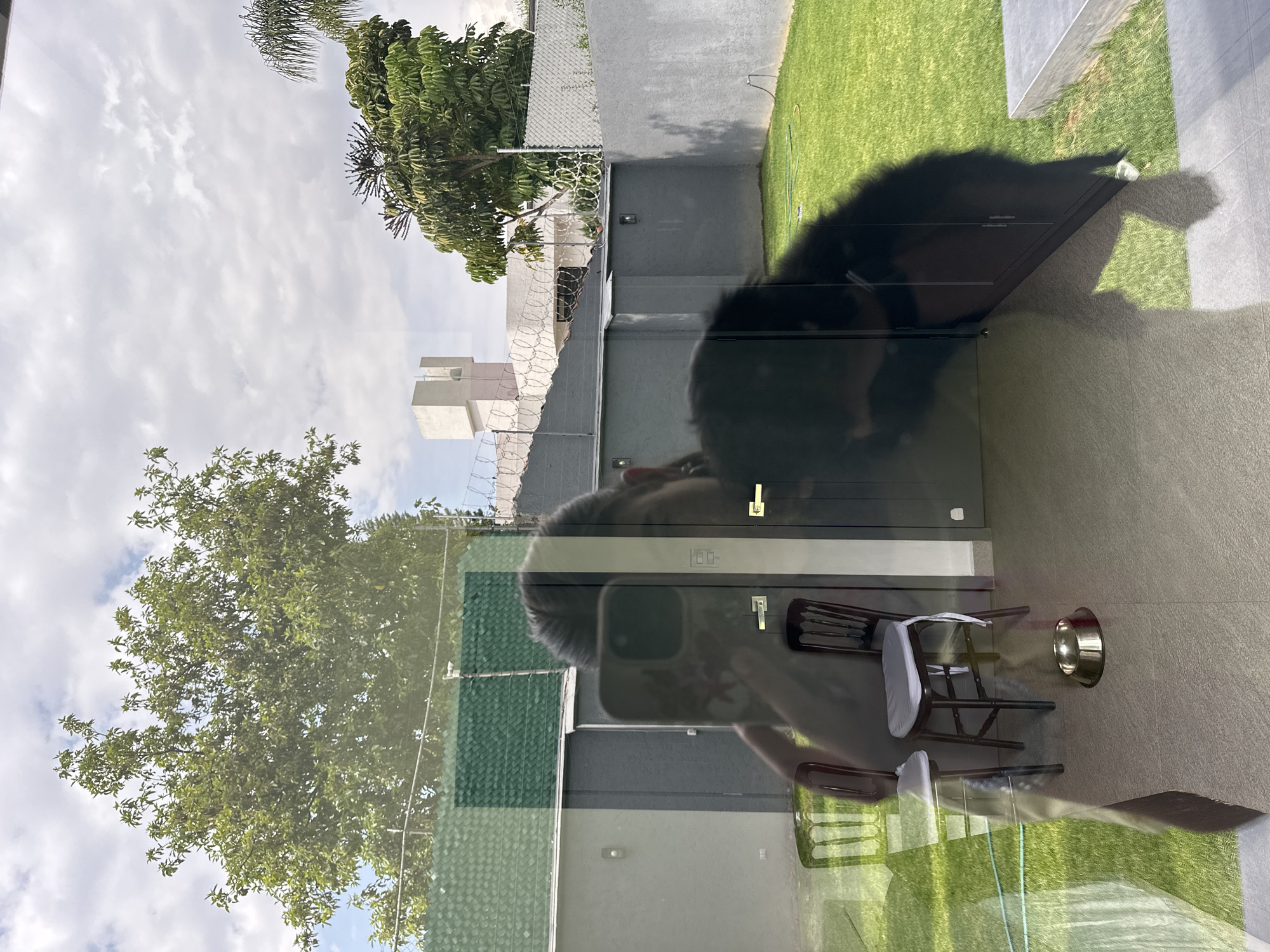 The image depicts a reflection in a window. The reflection includes trees, sky, a building with a fence topped with barbed wire, and a person holding a smartphone taking the picture. The foreground shows a patio area with a black surface, possibly a table, two chairs with white cushions, and a dog bowl. The location is in the city of Puebla, Mexico, but specific landmarks are not discernible. The focus is on the reflection and the items on the patio. The environment suggests an outdoor living space.