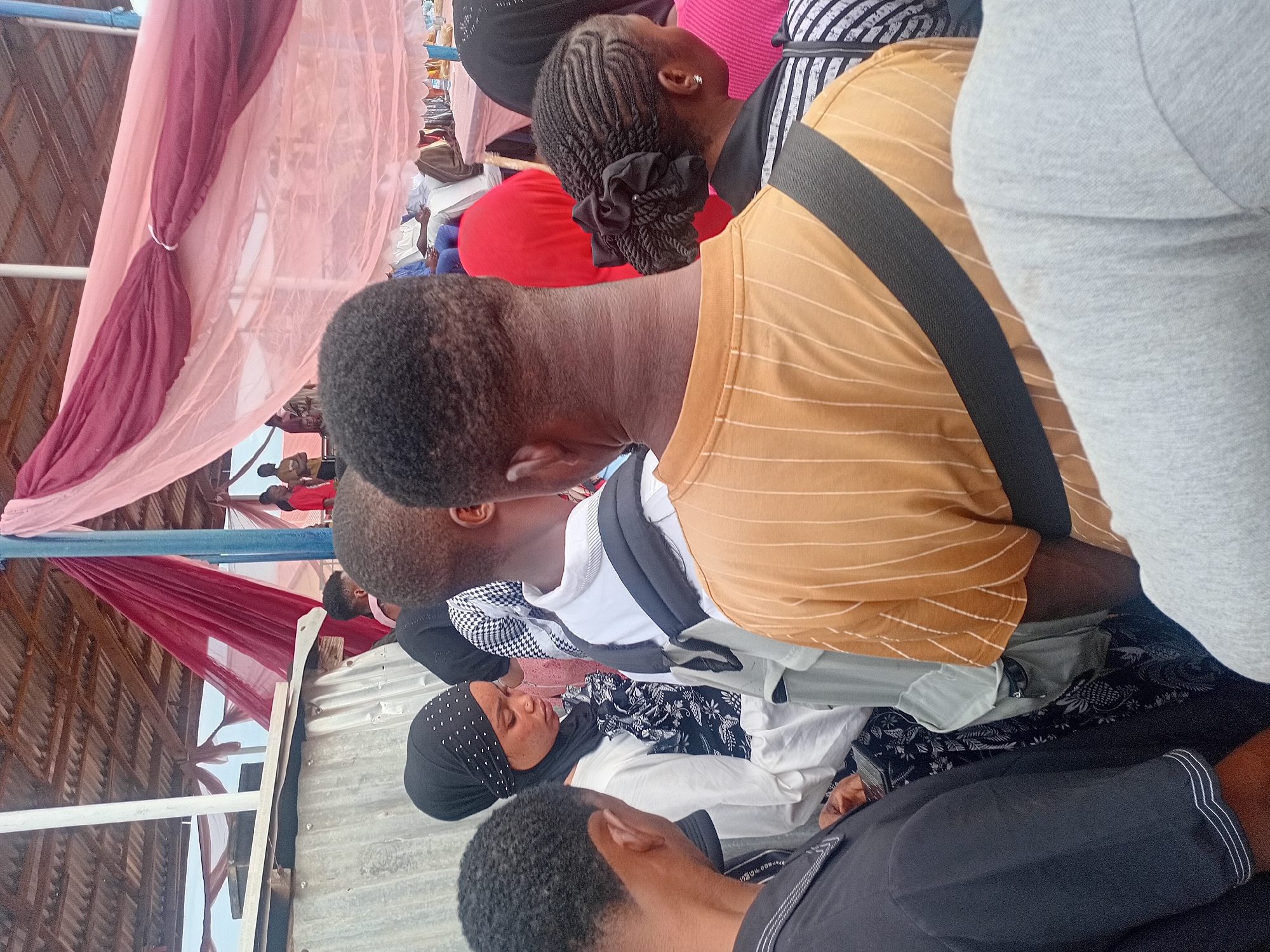 The image displays a crowded gathering occurring under a corrugated metal roof in Ogbogbo, Nigeria. Red and light pink sheer fabrics are draped and tied from the ceiling structure. On the far left, a corrugated metal partition stands next to a white vertical pole.
In the foreground, several individuals of African descent are standing. The leftmost person, viewed from behind, has short dark hair and wears a black collared shirt with white piping on the sleeves. Adjacent to them, a woman in a black hijab adorned with white dot embellishments and a white long-sleeved top over a dark patterned garment is looking downwards, revealing a nose piercing. To her right, another individual, seen from behind, wears a white long-sleeved shirt with a black and white patterned layer visible underneath. Further right, a person wears a mustard yellow short-sleeved t-shirt with vertical white stripes and has a black strap, possibly from a bag, across their shoulder. Partially visible behind this person is a woman with braided hair styled into a bun with a black tie, wearing a black and white striped top and an earring.
In the mid-ground and background, other attendees are seated on chairs, some dressed in traditional white robes and blue trousers. A table covered with a pink cloth is visible in the background. The individuals in the foreground are standing in close proximity, appearing to observe or wait.