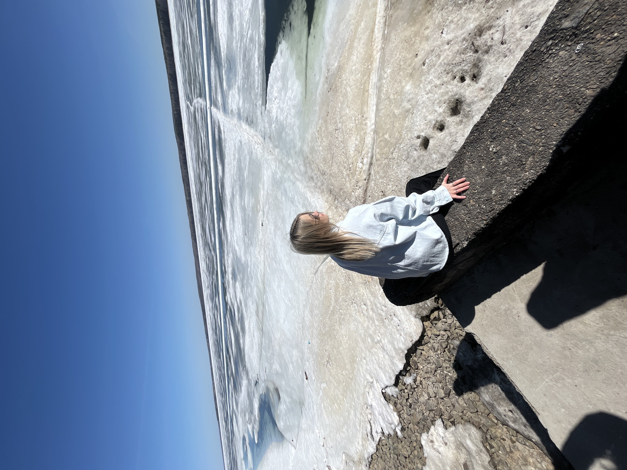 A woman with long blonde hair, wearing a light blue denim jacket, sits on a concrete edge overlooking a frozen body of water, possibly a river or lake, near Chaikovsky, Russia. The sky is clear blue. The edge where she sits is textured concrete, with some loose gravel. The ground leading to the frozen water is covered in snow and ice. The body of water is mostly frozen with patches of open water. Some trees are visible in the distance.