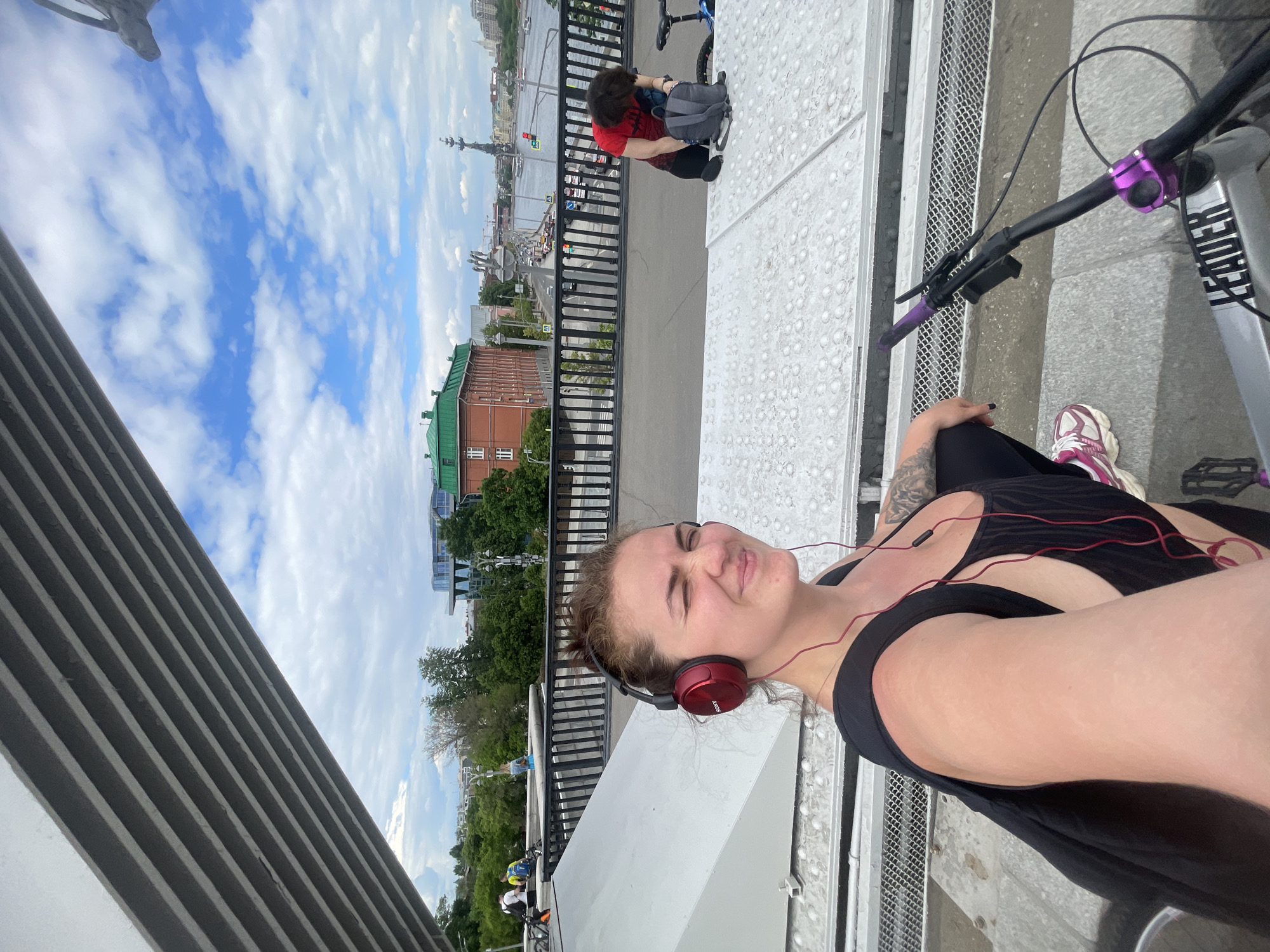 The image is a medium shot of a woman wearing headphones, with a cityscape in the background. She is on a bridge or elevated structure, with a bicycle partially visible in the frame. Another person is visible further down the structure, crouching beside a bicycle. The background includes a red brick building with a green roof, and a monument. The sky is blue with white clouds. The city of Moscow, Russia is identified as the location.