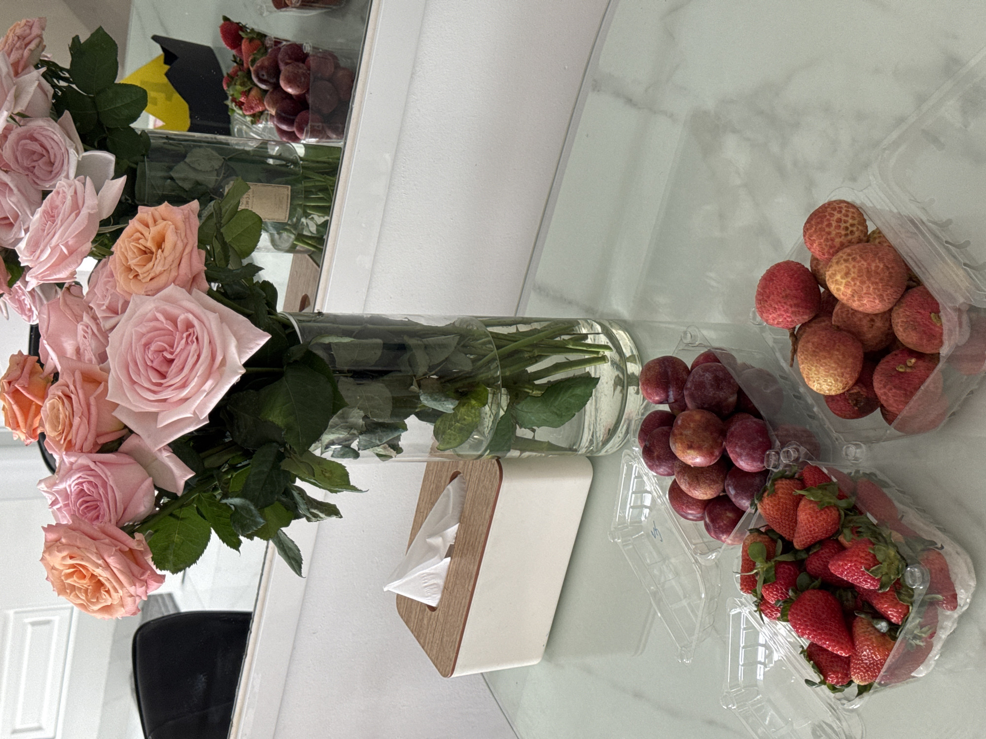 A slightly canted overhead shot reveals a tabletop arrangement, likely within a residence, potentially in Bac Nha Trang Ward, Vietnam, though this is not directly verifiable. On the left, a large vase filled with water and an assortment of pink and peach roses is visible. Next to the vase is a rectangular tissue box, white with a light wood trim, from which a tissue protrudes. To the right of these items are three clear plastic containers, each filled with different types of fruit: lychees, plums, and strawberries. The table's surface appears to be a light-colored marble or similar material with a subtle veining pattern. A black chair is partially visible in the lower-left corner. Other than these objects, there is no other sign of humans or other subjects.