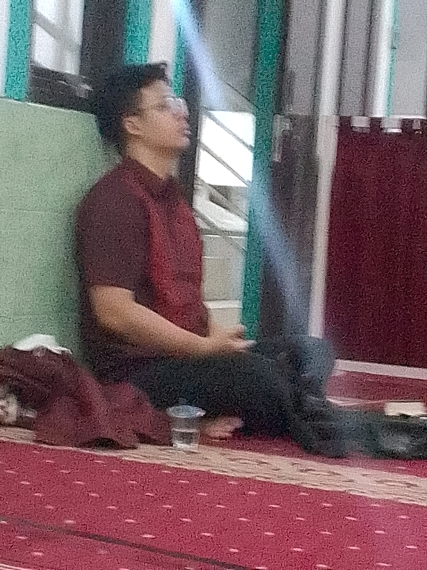 A young man with short, dark hair and glasses is sitting on a red carpeted floor, leaning against a light green wall. He is wearing a dark red polo shirt and dark pants. His head is tilted back, and his eyes are closed or looking upwards. To his left, a folded burgundy cloth lies on the floor. Further to the right, a clear glass with a small amount of liquid is visible on the floor. The background shows vertical structures with a dark red fabric hanging on the right and a white column. The image appears to be taken indoors, possibly in a place of worship or a community hall, given the carpeted floor and the overall decor. The lighting suggests a natural light source coming from the upper left.