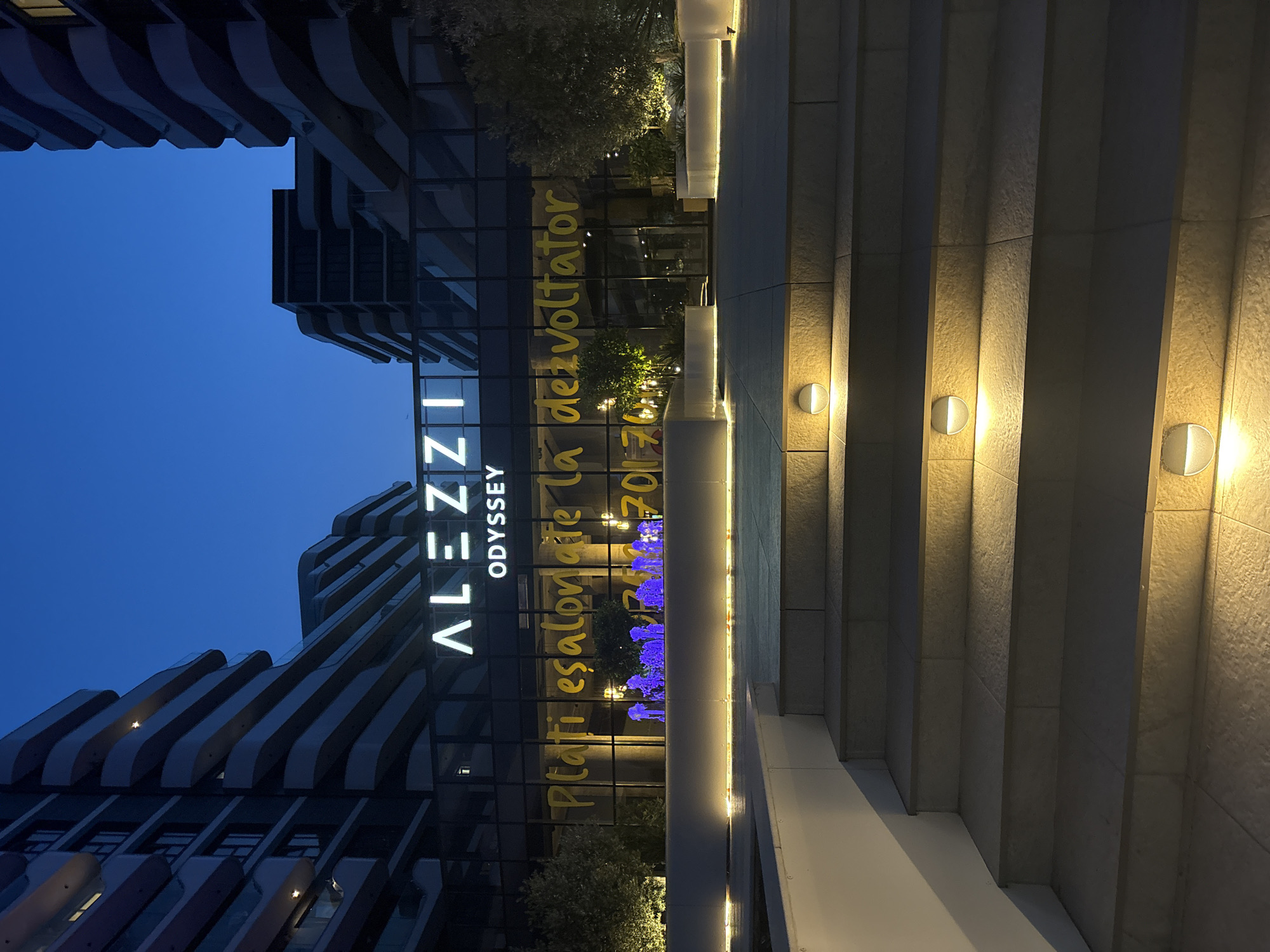 This is a vertical, low-angle shot of a modern building at dusk or night. The building features a distinctive undulating facade with ribbed elements and glass windows. On the glass facade, the illuminated white text "ALEZZI ODYSSEY" is visible. To the right of this, yellow text is painted or projected onto the glass, reading "Plătiți eșalonat la dezvoltator 201-70...". Blue lights are also visible behind the glass, illuminating some bushes.
In the foreground, a set of wide, illuminated concrete stairs leads upwards. Wall-mounted lights with a warm glow are spaced along the stairs and the adjacent textured concrete wall. The sky is a deep, clear blue, indicating either twilight or nighttime. The overall impression is one of modern architecture and urban development, possibly a residential or commercial complex. The scene conveys a sense of calm and sophistication, with the artificial lighting highlighting the architectural features. The text suggests a real estate development or sales promotion. The location is identified as Năvodari, Romania.