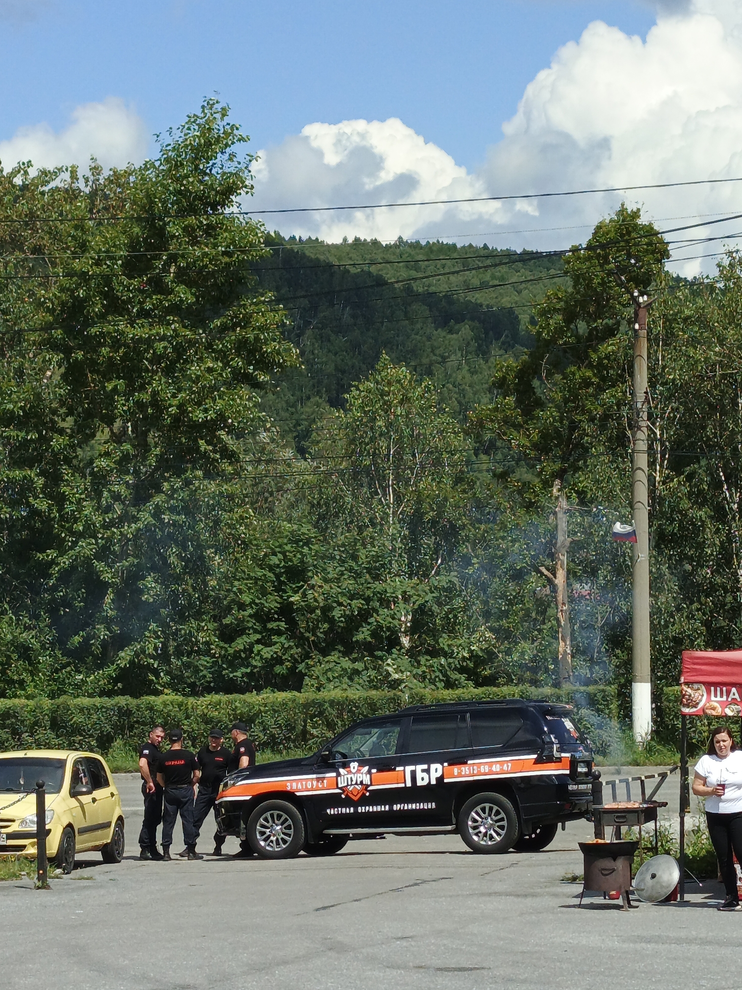 In Zlatoust, Russia, on a sunny day with scattered clouds, a scene unfolds in an outdoor area with dense green trees and a hilly backdrop. Four men in black uniforms with the inscription "ОХРАНА" (Okhrana - Security) on their backs stand in a huddle near a yellow car. Adjacent to them is a black SUV with an orange stripe and Cyrillic lettering, including "ГБР" (GBR) and a phone number. The SUV appears to be a security vehicle. Further to the right, a woman in a white t-shirt and black pants stands near a food stall with a red awning, holding a drink. In front of the stall, a portable grill with food cooking and a large pot are visible. Smoke emanates from a fire pit or grill, contributing to the atmosphere. A utility pole with electrical wires and a small Russian flag attached is also present. The ground is paved, suggesting a parking lot or open area.