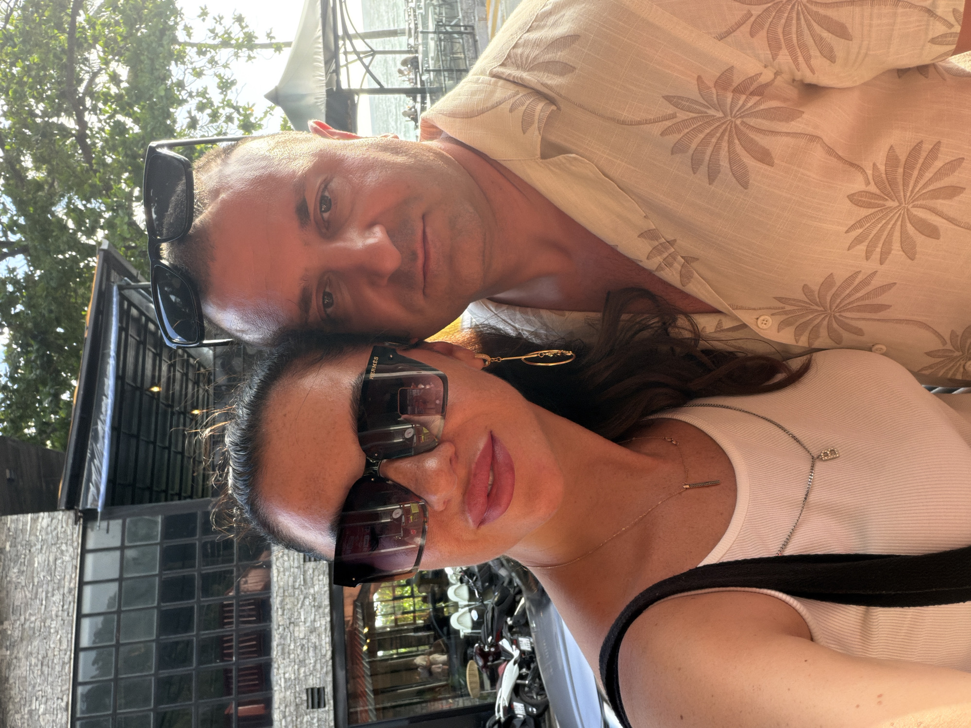 In Patong, Thailand, a couple poses for a selfie. The woman, in the foreground, wears large, dark sunglasses and a white tank top. She has a delicate gold necklace with a vertical pendant and another silver chain with a charm. Her lips are painted pink. The man, positioned behind her and slightly to the right, has sunglasses perched on his head and wears a light-colored short-sleeved shirt with a palm tree print. The background reveals a building with a stone facade and large windows, suggesting a restaurant or shop. Beyond that, trees and a glimpse of the sea with distant patrons at outdoor tables indicate a seaside location, likely a beachfront establishment in Patong. The overall lighting suggests it is either late afternoon or early morning.