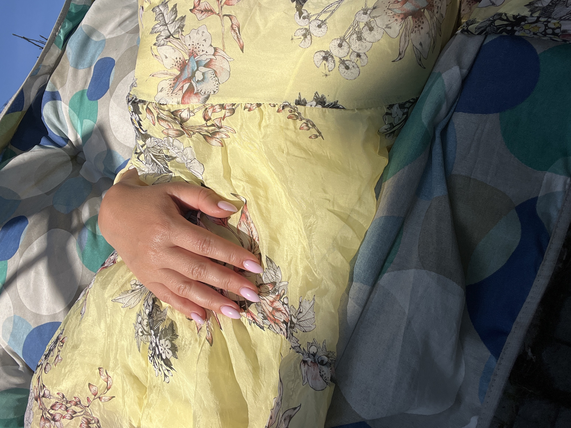 A close-up shot focuses on a person's hand resting on yellow fabric with a floral pattern. The hand is manicured with light pink polish. The fabric appears to be part of a garment or bedding. In the background, other fabrics with abstract circular patterns in shades of blue, green, and gray are visible. The scene is illuminated by natural light, casting shadows. The image was taken outdoors, with a clear blue sky visible at the top right. The location is specified as Роса Снт, Russia.