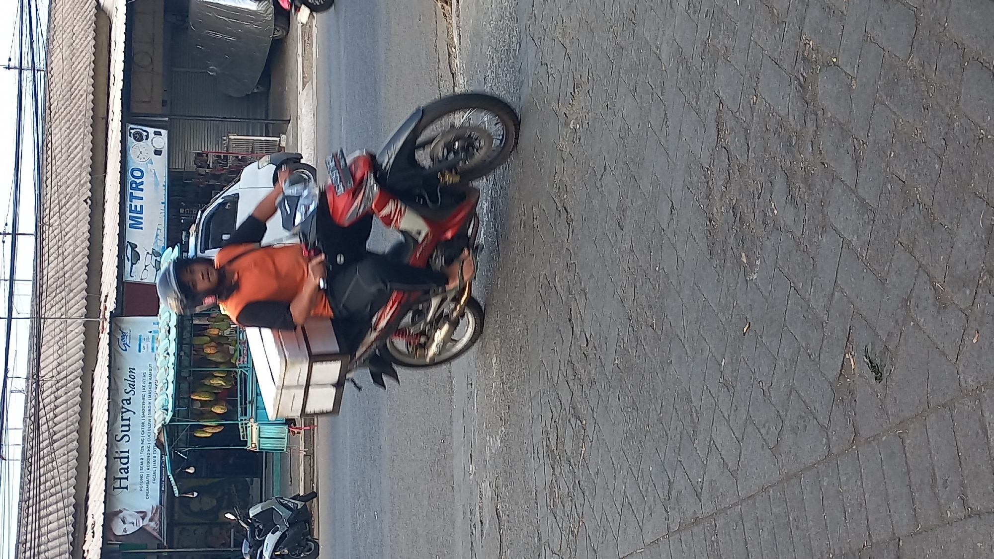 In Mangunsari, Indonesia, a man wearing an orange and black shirt and a black helmet rides a red and white motorcycle with a white cargo box on the back. He is on a street with gray, interlocking brick pavement. In the background, there are several buildings with corrugated metal roofs. One building has a banner that says "Hadi Surya Salon" and another banner reads "METRO". There is a white car and a covered vehicle parked near the buildings. A scooter is parked on the left side of the street.