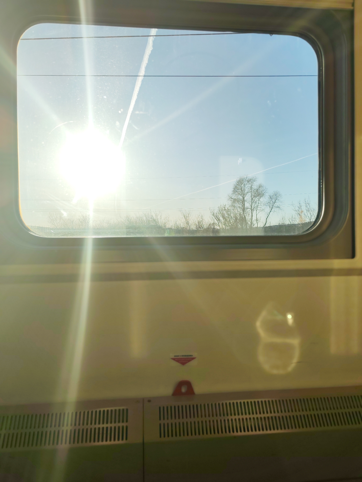 The image displays an exterior view from inside a train, seen through a rounded rectangular window with a silver-grey frame. The scene outside features a clear blue sky dominated by a brilliant, low-angled sun on the left, creating significant glare and lens flares across the window and the train's interior panel. Several thin white contrails are visible in the sky, one cutting through the sun's brightness. Horizontal power lines or wires stretch across the view, with a line of bare trees and bushes, devoid of leaves, visible below the horizon, suggesting winter or early spring.
The lower portion of the image shows the train's interior wall, which is a light beige or yellow. Bright sun rays fall upon this panel, creating vertical streaks of light. Towards the bottom, ventilation grilles are visible. A small red triangular pull handle or emergency release is present below the window, next to a small red triangular sticker. This sticker bears the visible text "UWAGA!", which is Polish for "ATTENTION!". A large, bright, and distorted reflection, somewhat resembling a rounded figure '8', is prominently cast onto the interior panel to the right. The image captures a sunny daytime train journey through a natural landscape. No people are visible.