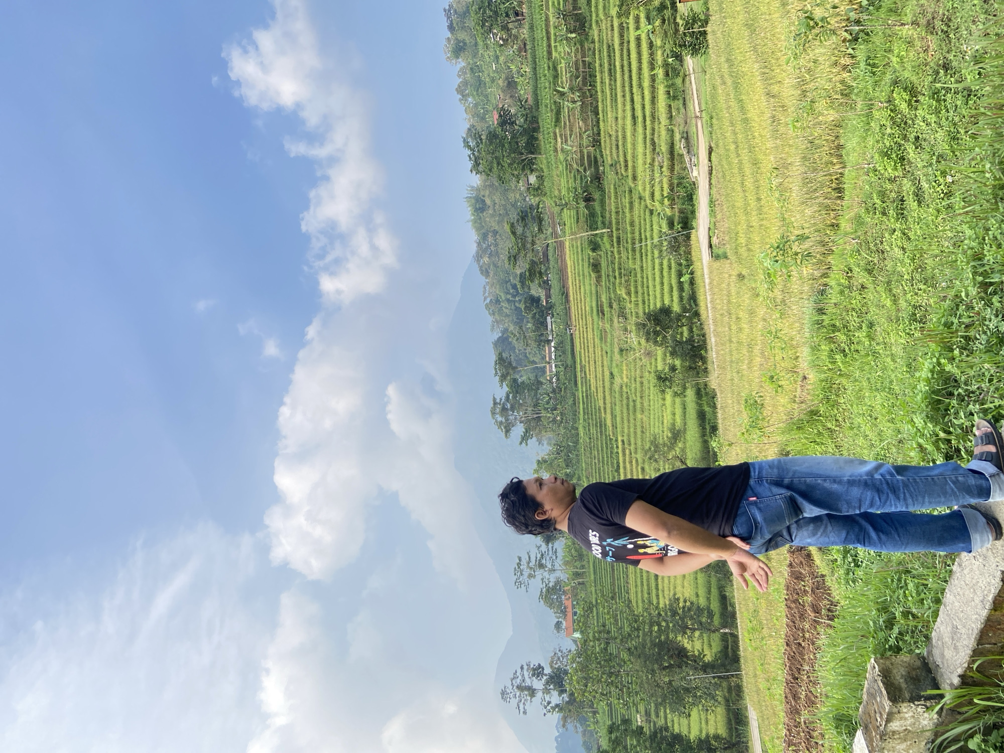 A person wearing a black T-shirt and blue jeans is standing in a rural area of Penanggungan, Indonesia. To the person's right is a green hillside area, and a rice paddy is visible in the background. Above, the sky is blue with white clouds. The person appears to be looking upwards. The image is oriented sideways.