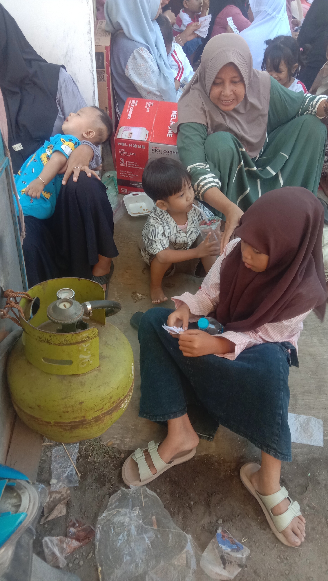 The image captures a scene in the city of Karang Anyar, Indonesia, showing a group of people, primarily women and children, gathered outdoors. A large yellow LPG gas cylinder is prominently positioned in the foreground, with a gas stove regulator attached. To the left, a baby wearing a blue and yellow giraffe-patterned outfit is being held and appears to be sleeping. Beside the baby, a woman in a black headscarf and dark skirt is seated.
In the middle ground, a young child in a striped shirt is sitting and drinking from a cup with a straw. Behind this child, an adult woman wearing a green headscarf and green dress is interacting with the children, appearing to hand something to one of them. A red box, possibly a rice cooker, is visible between the adults and the children.
To the right, a young girl wearing a brown headscarf and a light pink striped shirt is seated on the ground, holding a piece of paper and a small blue-capped bottle. She is wearing dark blue jeans and light-colored sandals. Another woman in dark jeans and light-colored sandals is seated to her right. The ground appears to be dusty and uneven, with some plastic debris scattered around. The overall setting suggests a casual outdoor gathering, possibly in a residential area or a public space.